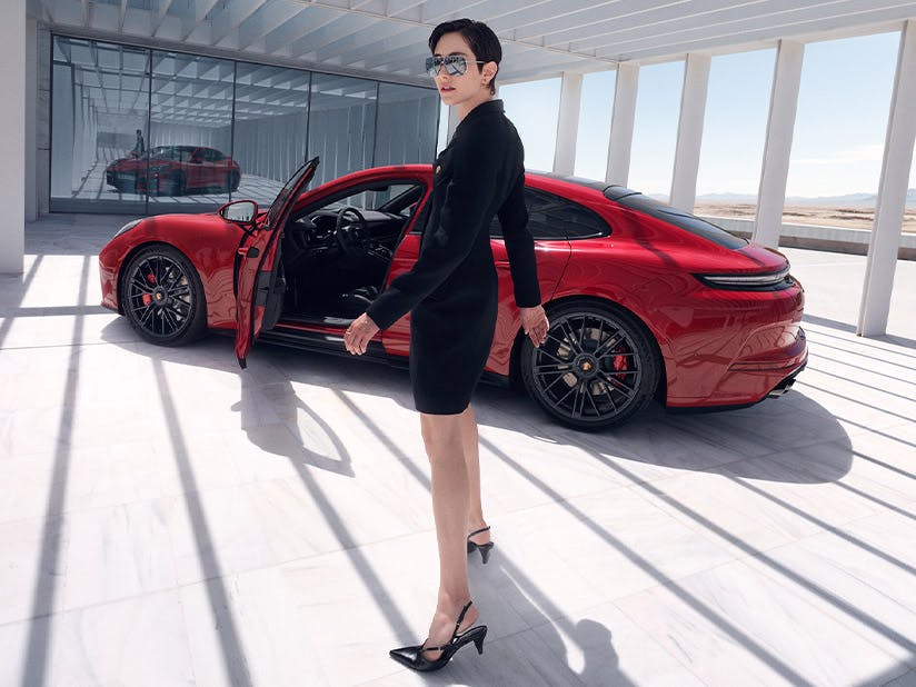 Person steht vor einem hellen Gebäude. Dahinter steht ein roter Porsche Panamera mit geöffneter Fahrertür.