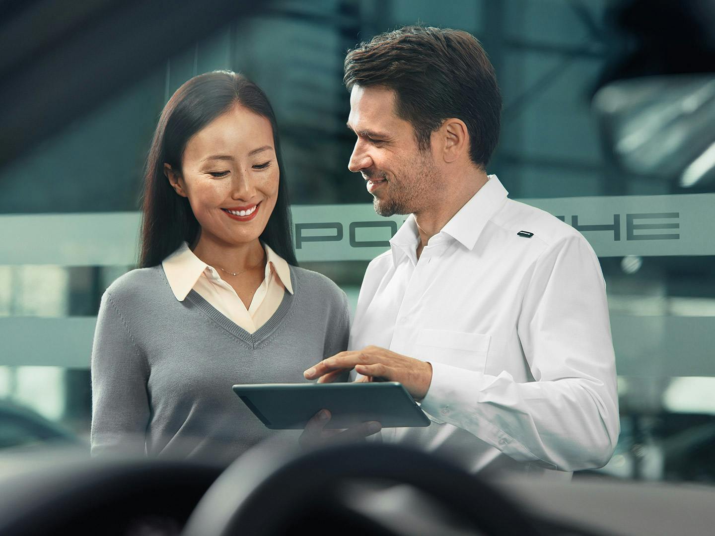 Durch die Scheibe eines Autos werden Porsche-Mitarbeiter und eine Kundin in einem Porsche-Showroom gezeigt. Der Mitarbeiter hält ein Tablet in der Hand.