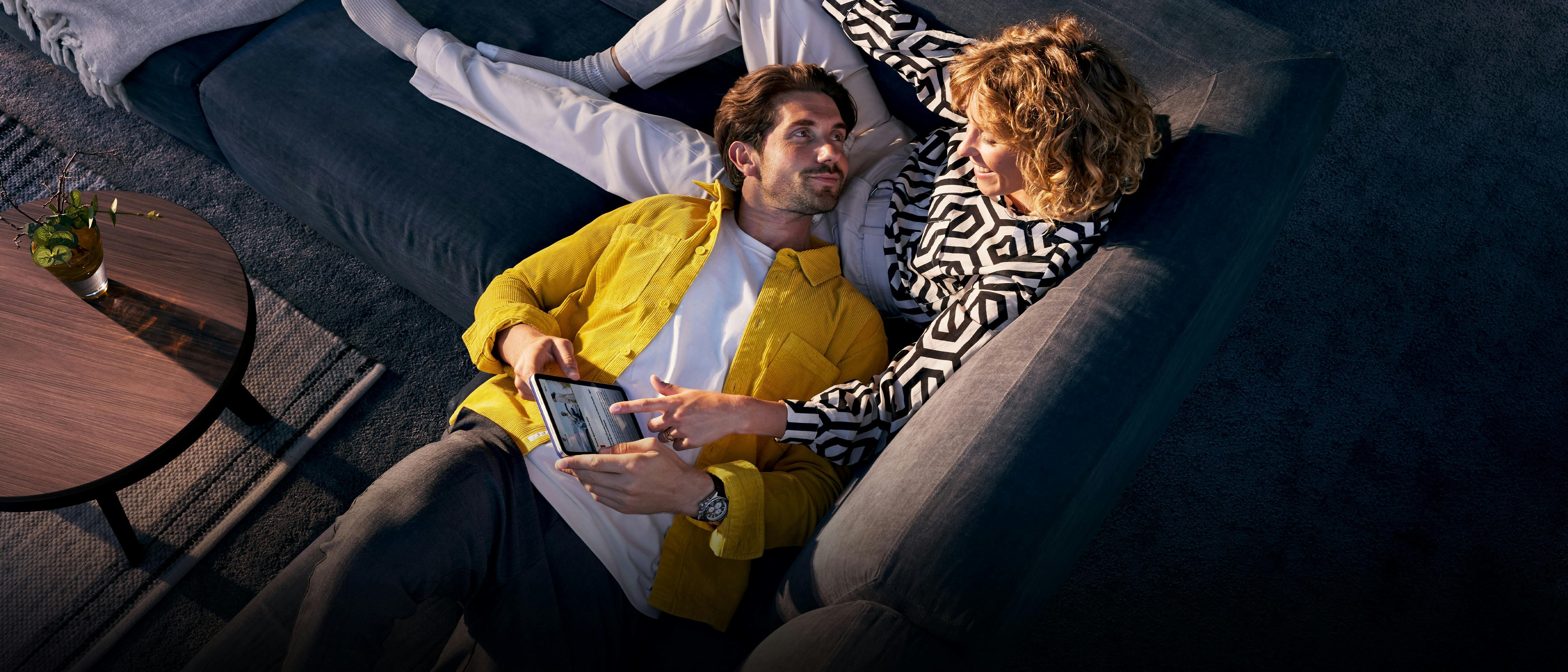 A couple are lying on the sofa. The woman is pointing at the screen of the tablet that the man is holding.
