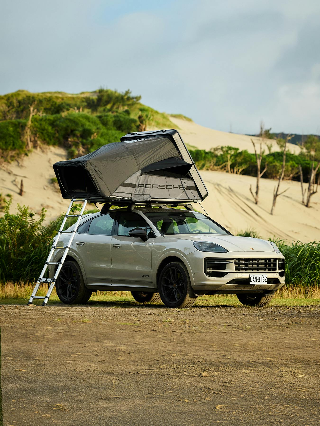A beige Porsche Cayenne with a rooftop tent and ladder parked on sandy terrain near dunes and greenery.