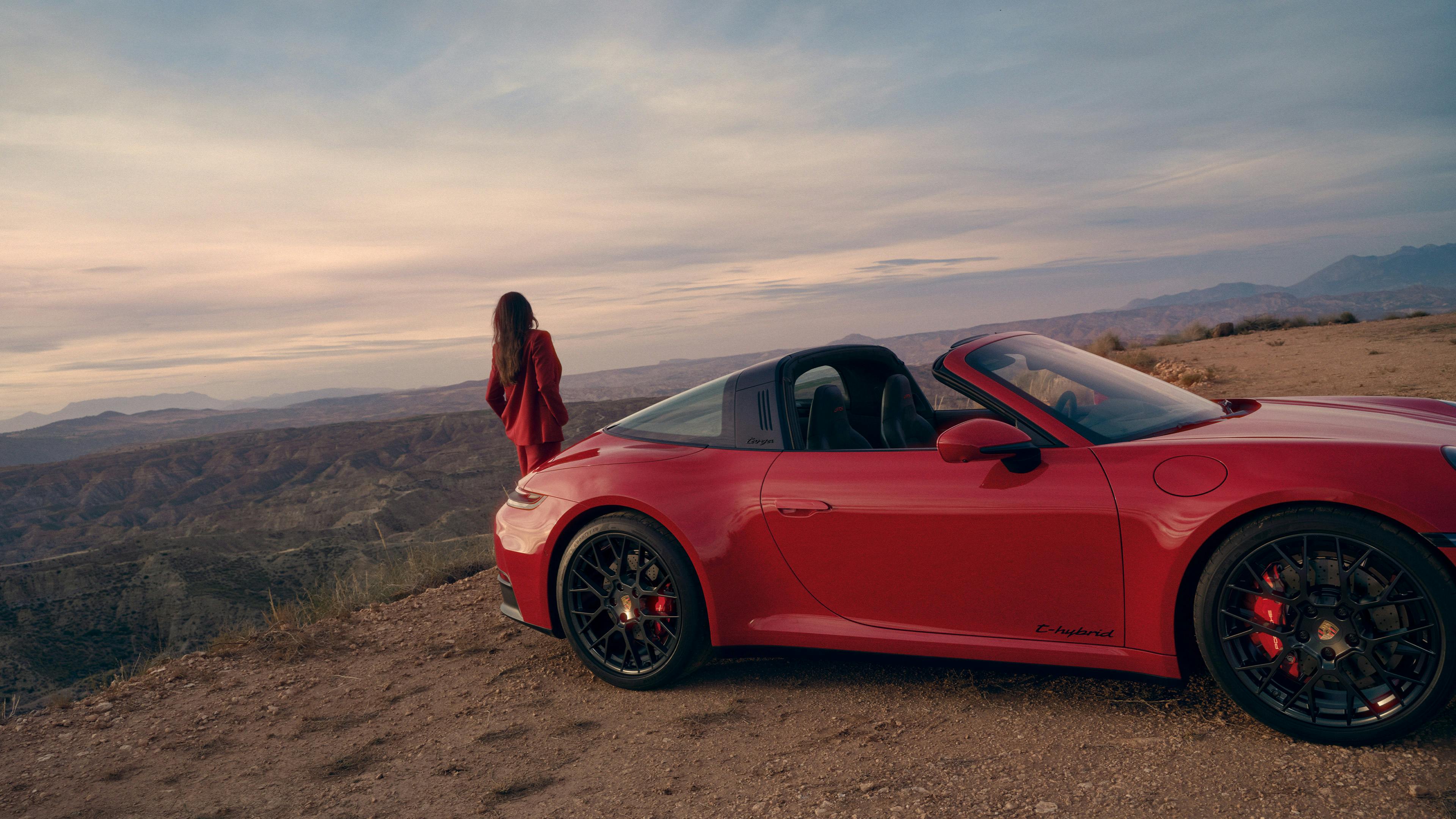 Roter Porsche 911 Targa steht auf einer Anhöhe in einer weiten Landschaft, während eine Person daneben steht und in die Ferne blickt.