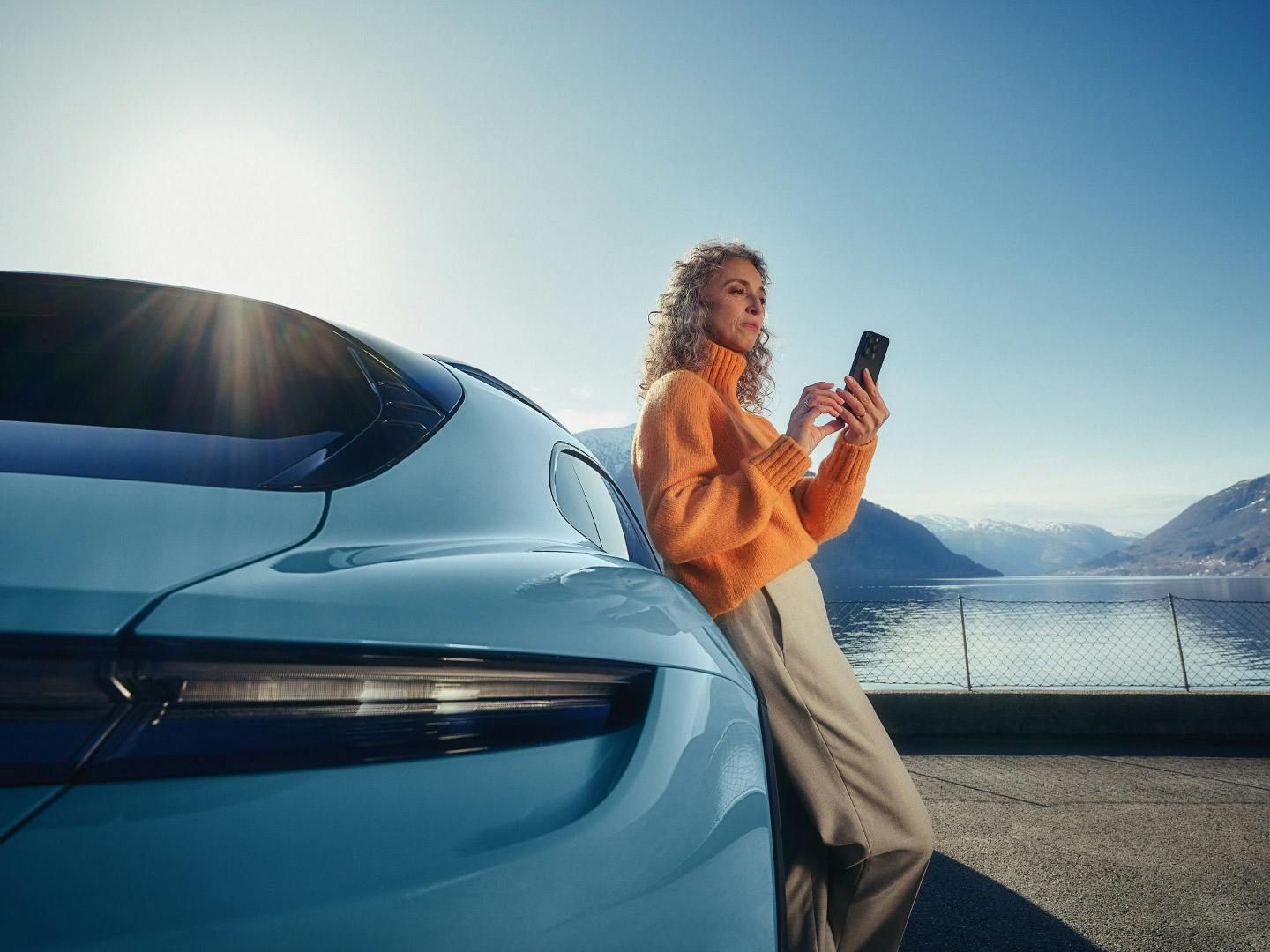 A woman looking at her phone while leaning against a parked light blue Porsche Taycan.