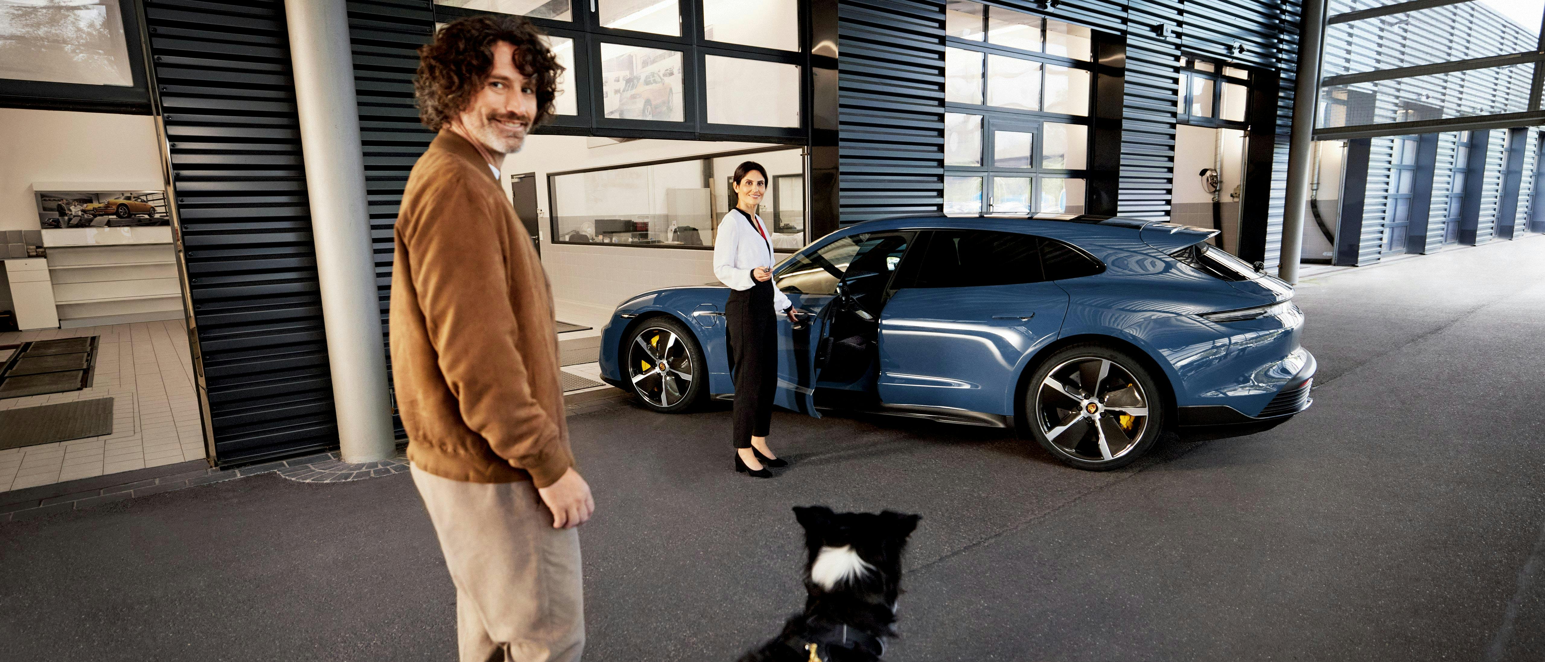 A man with a moustache looks into the camera, a dog stands in front of him. A blue Taycan can be seen in the background, its door being held open by a Porsche employee.