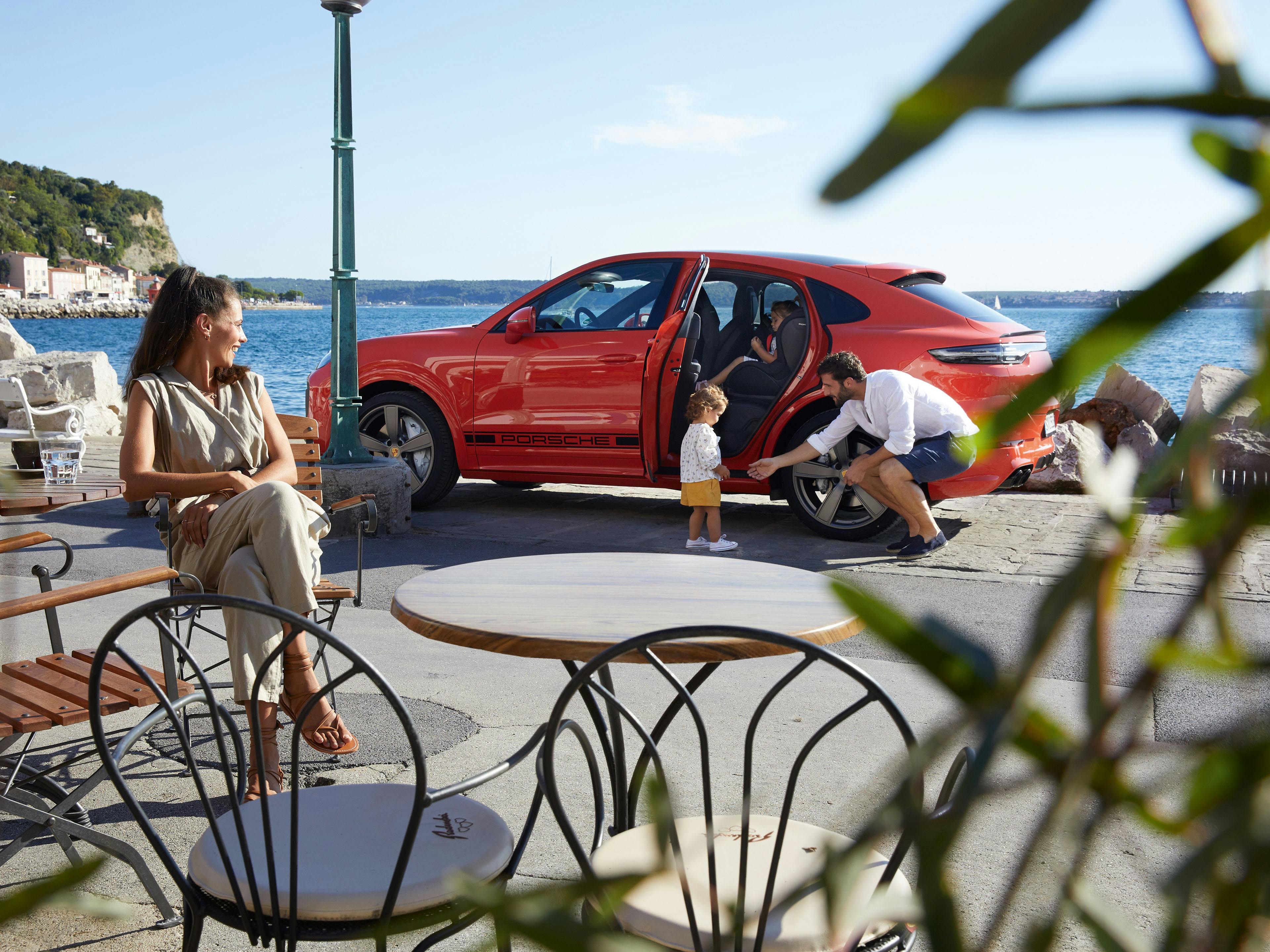 Ein roter Cayenne Turbo Coupé parkt an der Promenade, mit Vater, Kind davor und Frau im Cafe sitzend.
