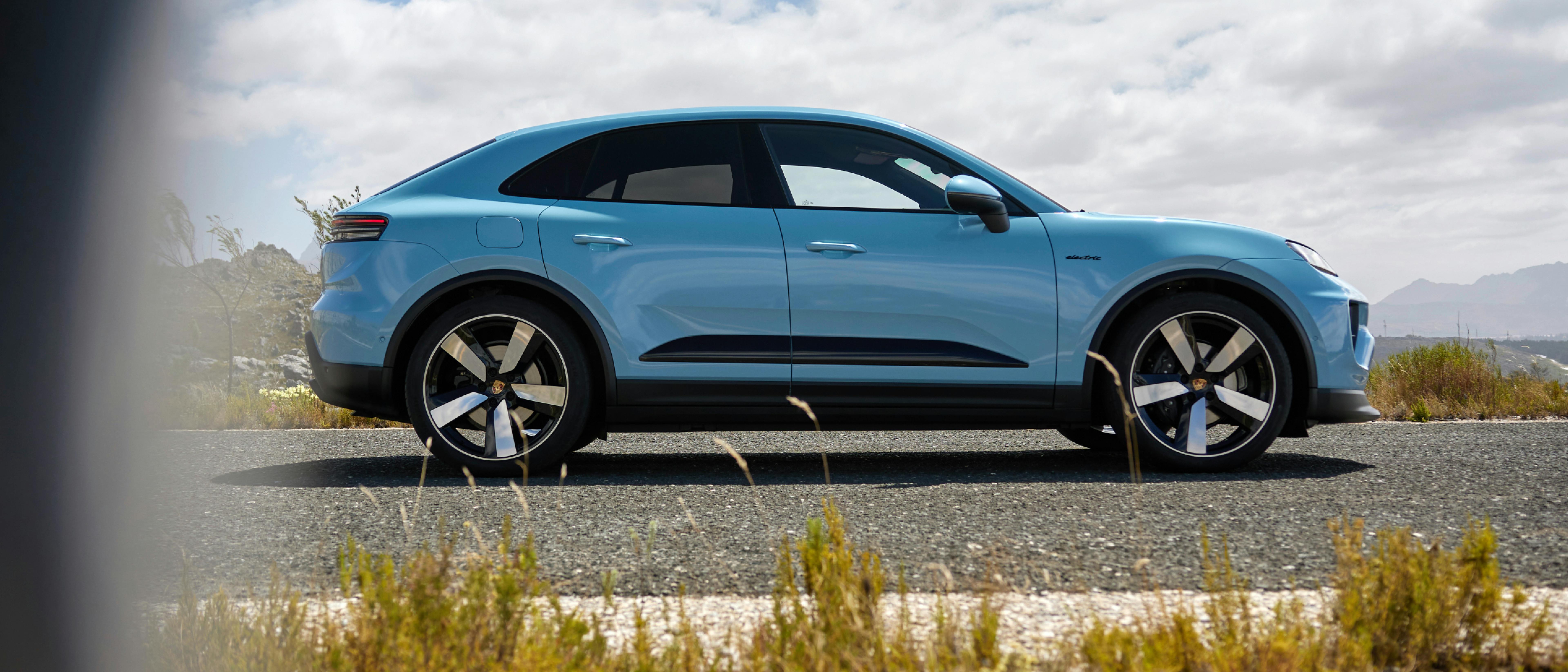 Porsche Macan in Frozen Blue Metallic parked on a road in a natural landscape with a Blue, slightly cloudy sky. Side view of the vehicle.