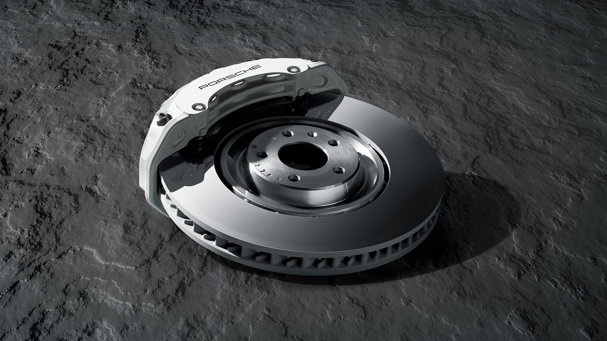 A white Porsche brake calliper and a perforated brake disc lie on a textured concrete slab.