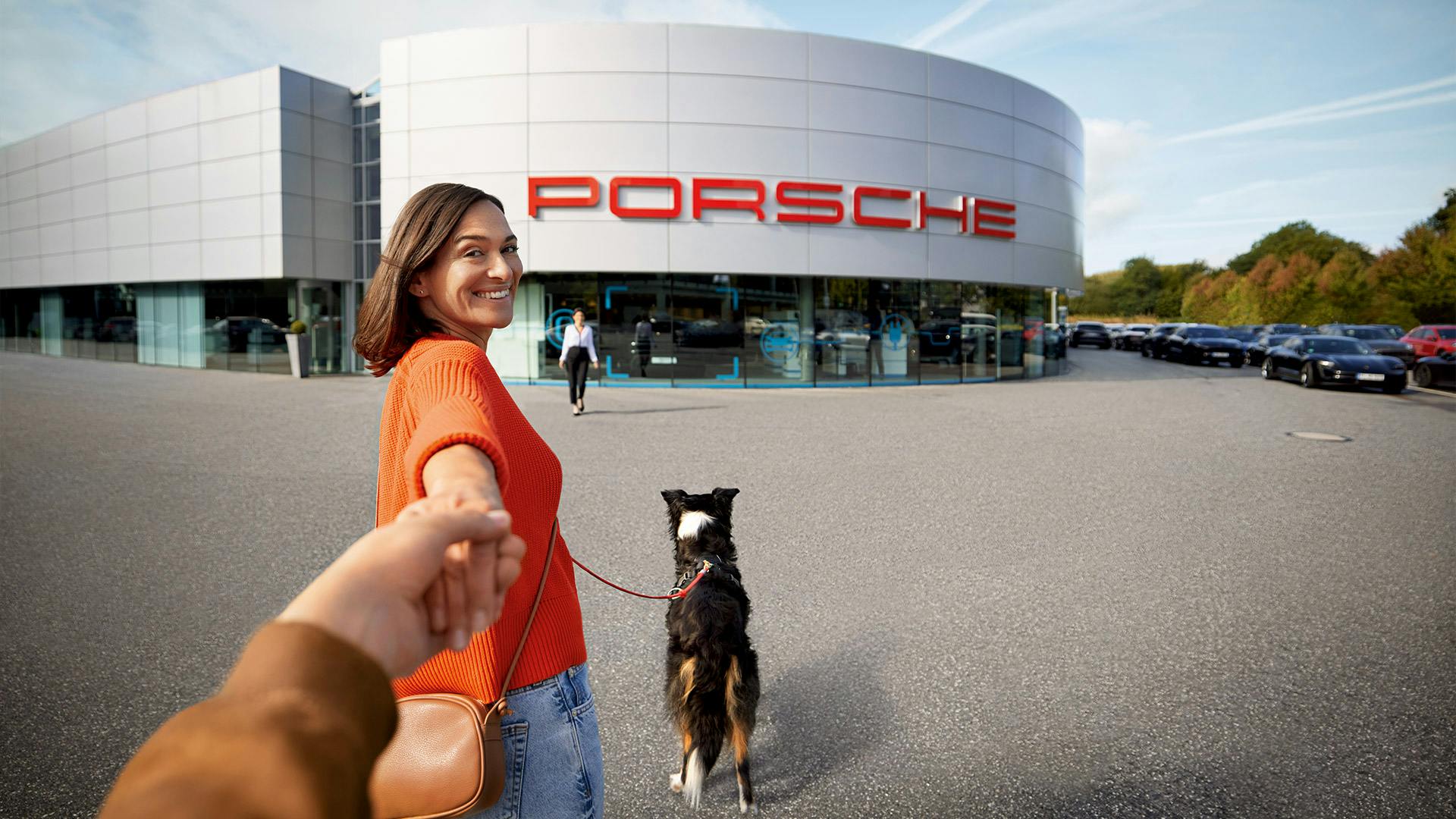 Eine  Frau in einem orangefarbenem T-Shirt, spaziert mit ihrem Hund auf ein Porsche Autohauses zu.
