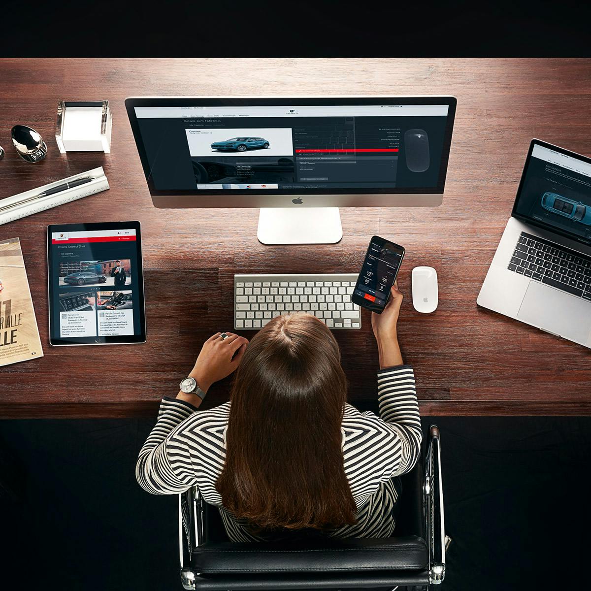Une table de travail est photographiée d'en haut. Un iMac, un MacBook et un iPad sont visibles sur la table. Une femme est assise à la table et tient son téléphone portable à la main. L'application My Porsche est ouverte sur tous les appareils.