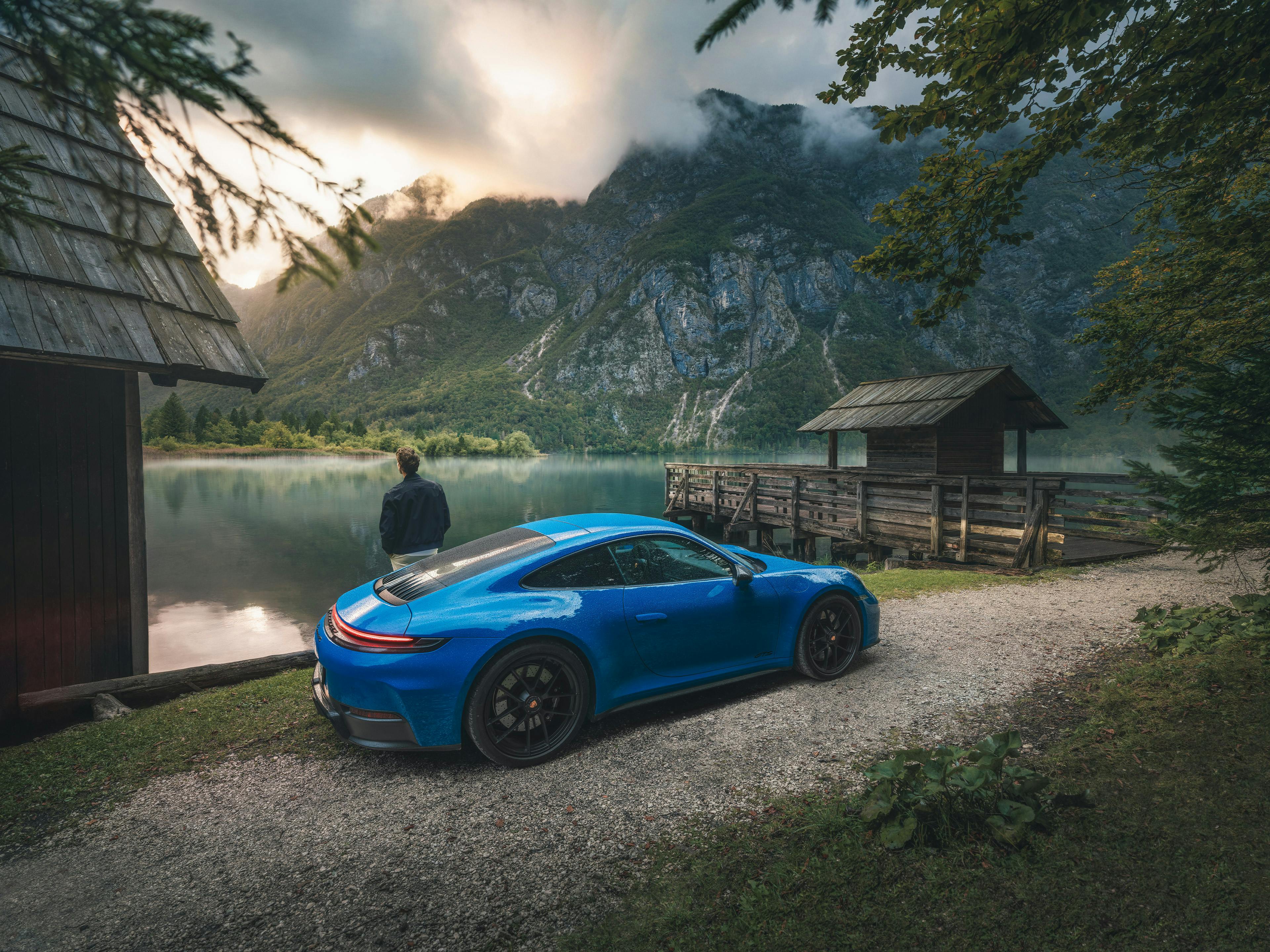 Un homme appuyé contre une Porsche 911 bleue stationnée devant un chalet au bord d’un lac, avec des montagnes en arrière-plan.