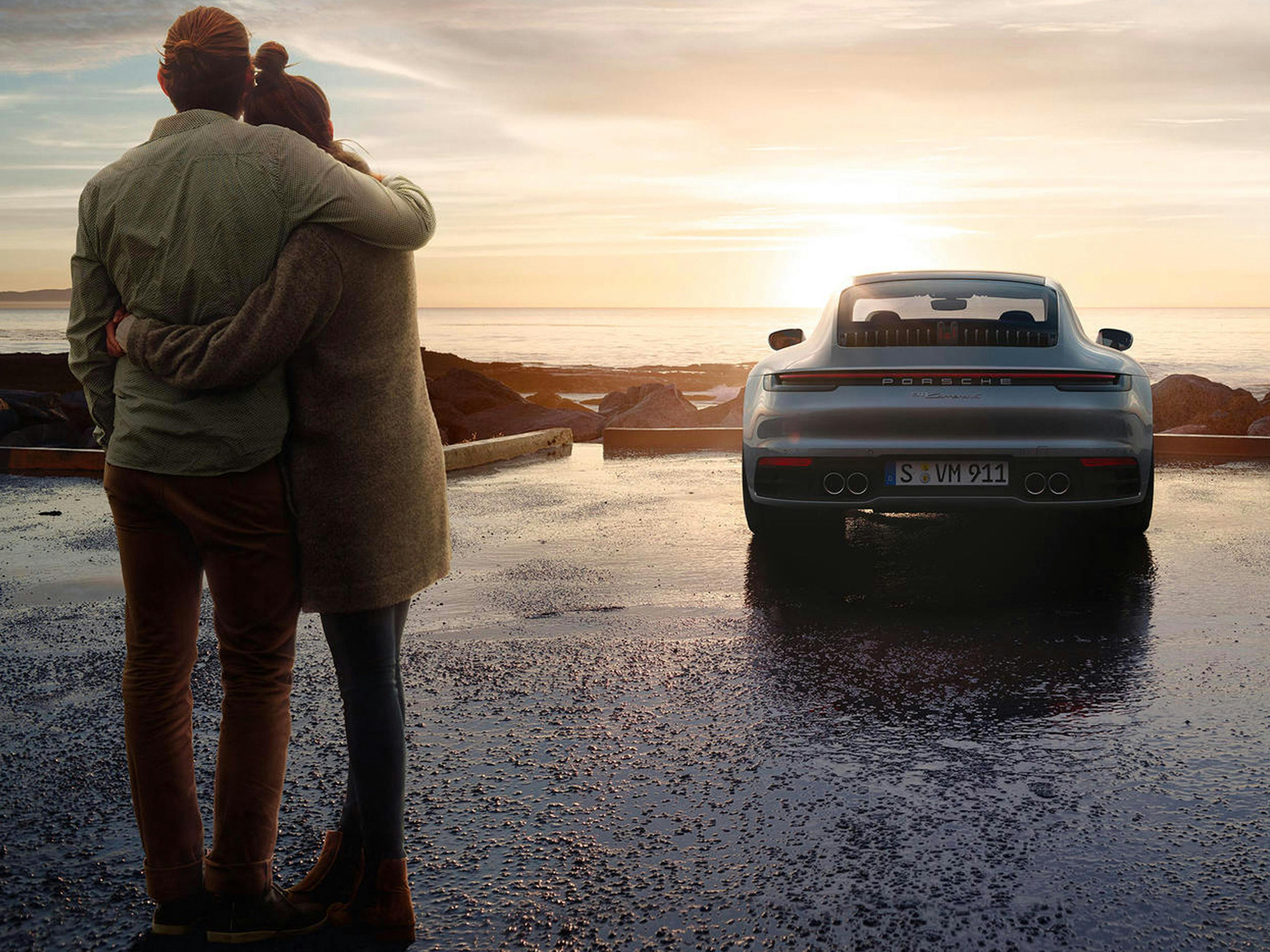 Una coppia si abbraccia comodamente al tramonto in riva al mare davanti a una Porsche 911, mentre sullo sfondo si vede il mare calmo.