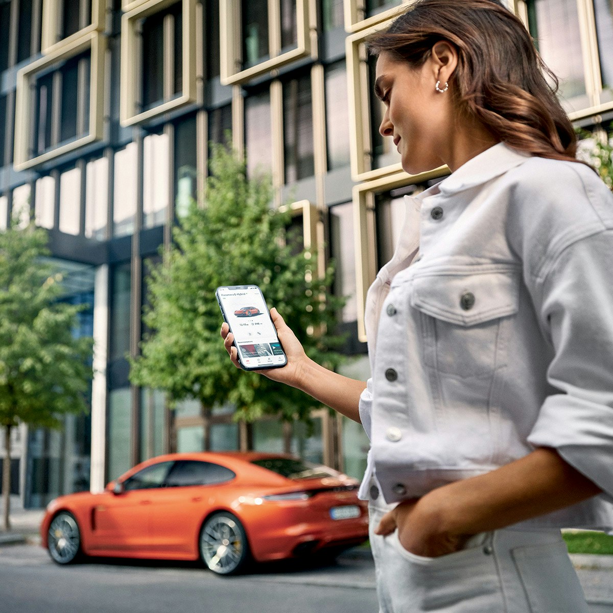 A young woman in the street, looking into a smartphone that is holding in her hand and a Porsche in the background.