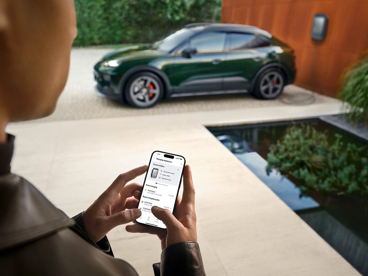 A person looking at their smartphone. A Porsche Macan 4S is charging at a Wallbox in the background.