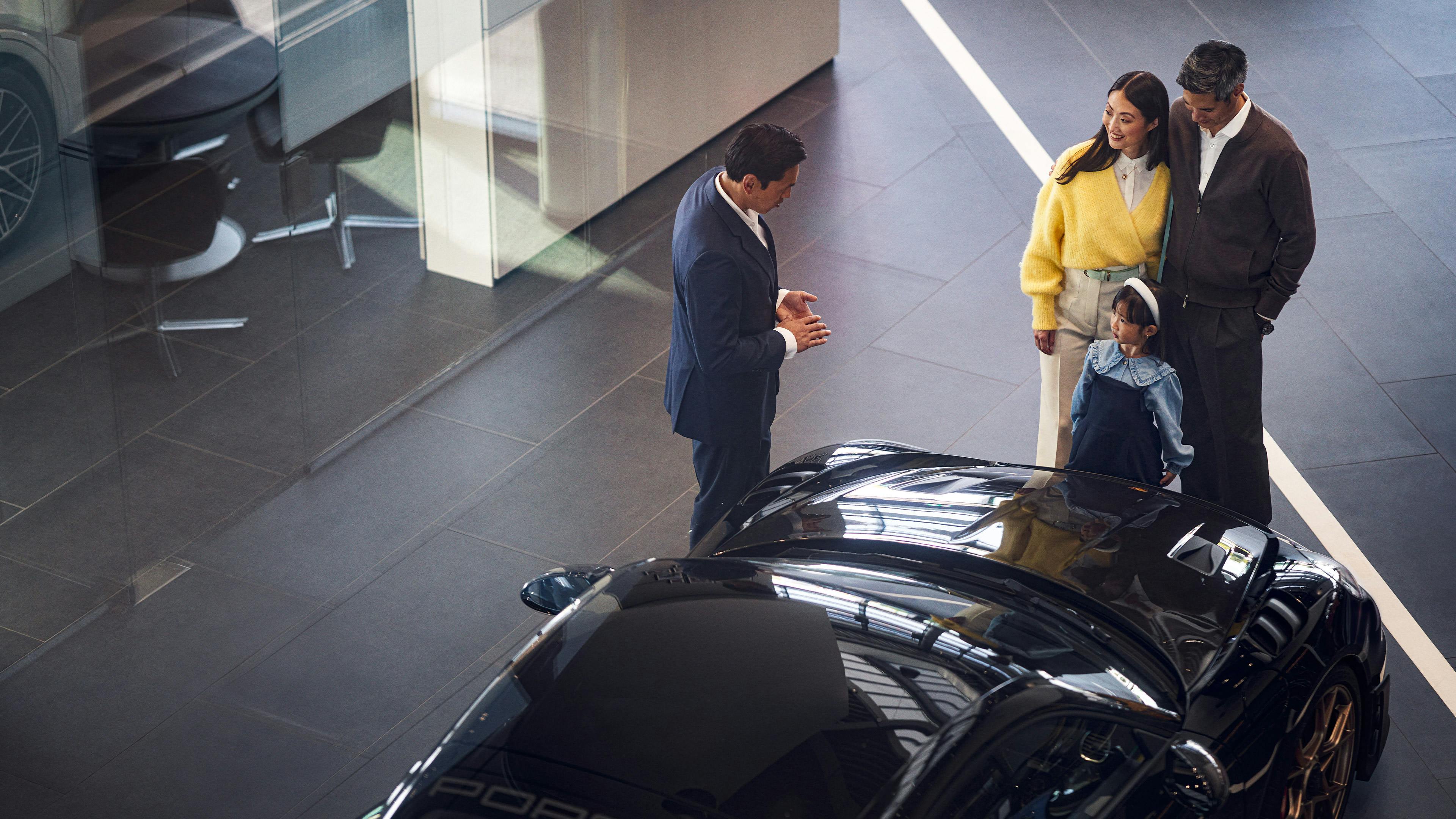 A bird's eye view shows a customer situation: a couple and a Porsche consultant are in the Porsche showroom. A Porsche Taycan can be seen in the background.
