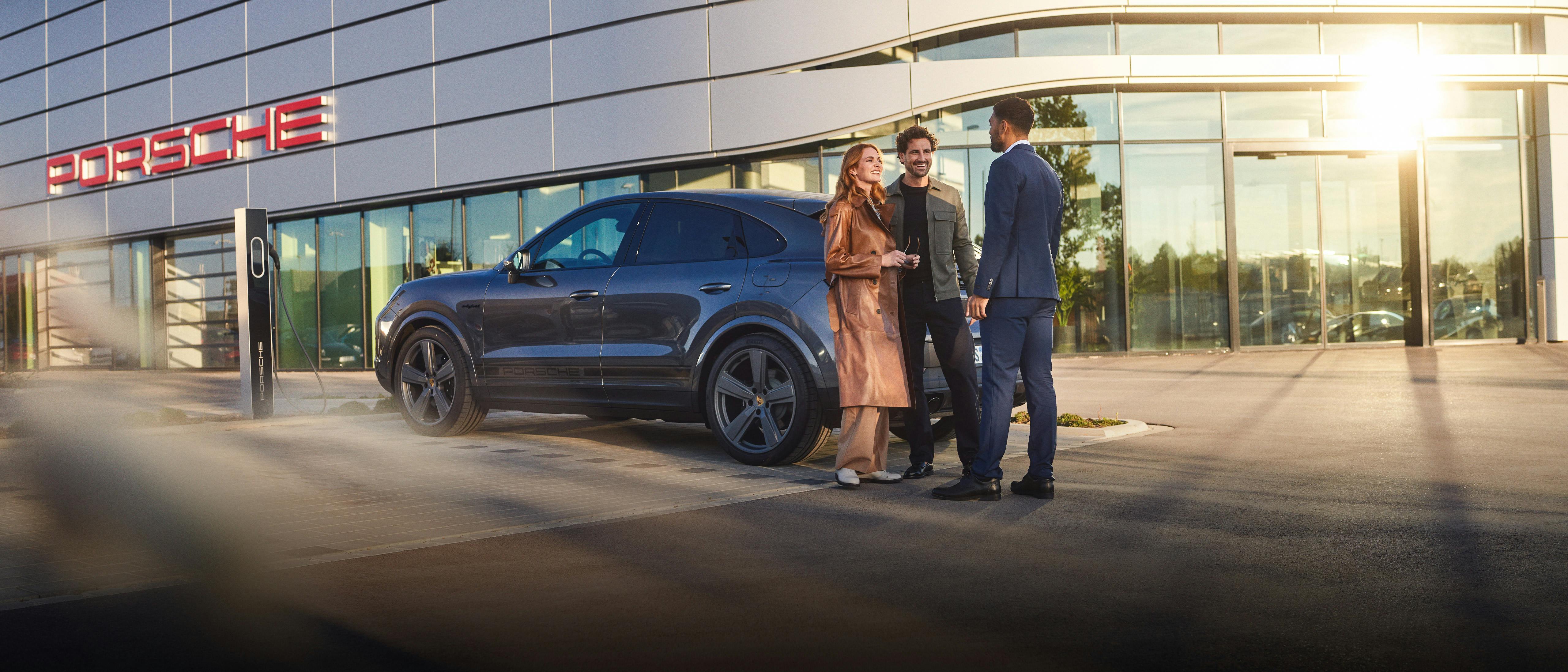 Un couple et un conseiller du Centre Porsche restent ensemble près d'un véhicule Porsche devant l'entrée du Centre Porsche.