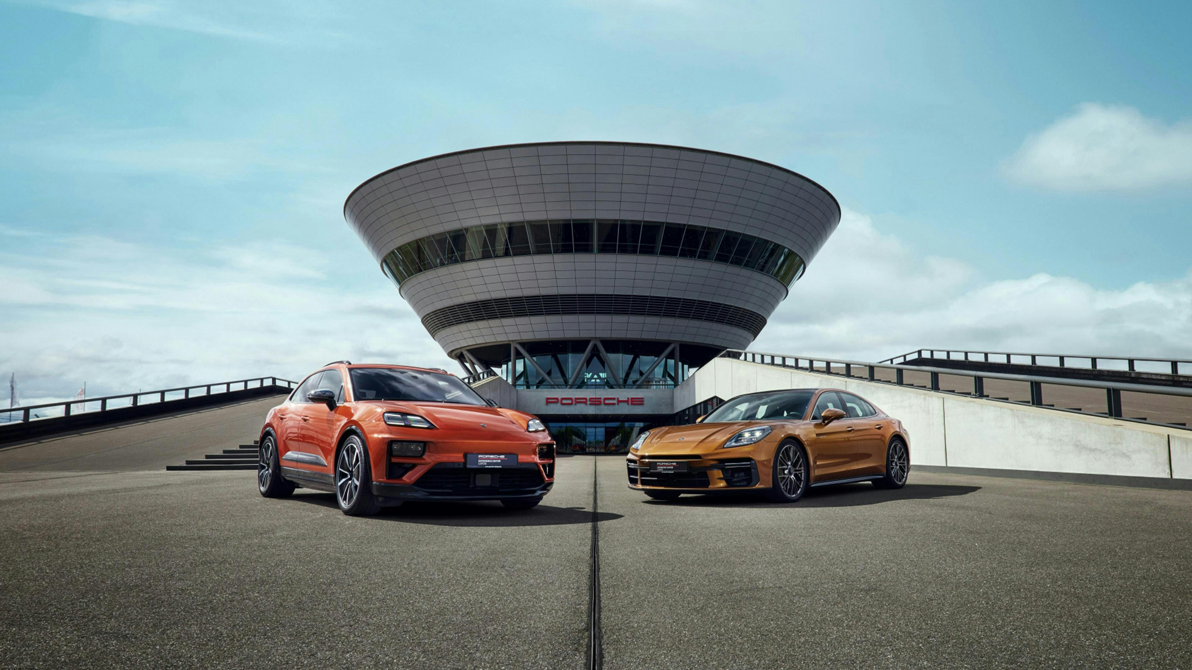 Ein orangefarbener Macan 4 und eine kupferfarbene Panamera, stehen nebeneinander vor dem Porsche Experience Center Leipzig.