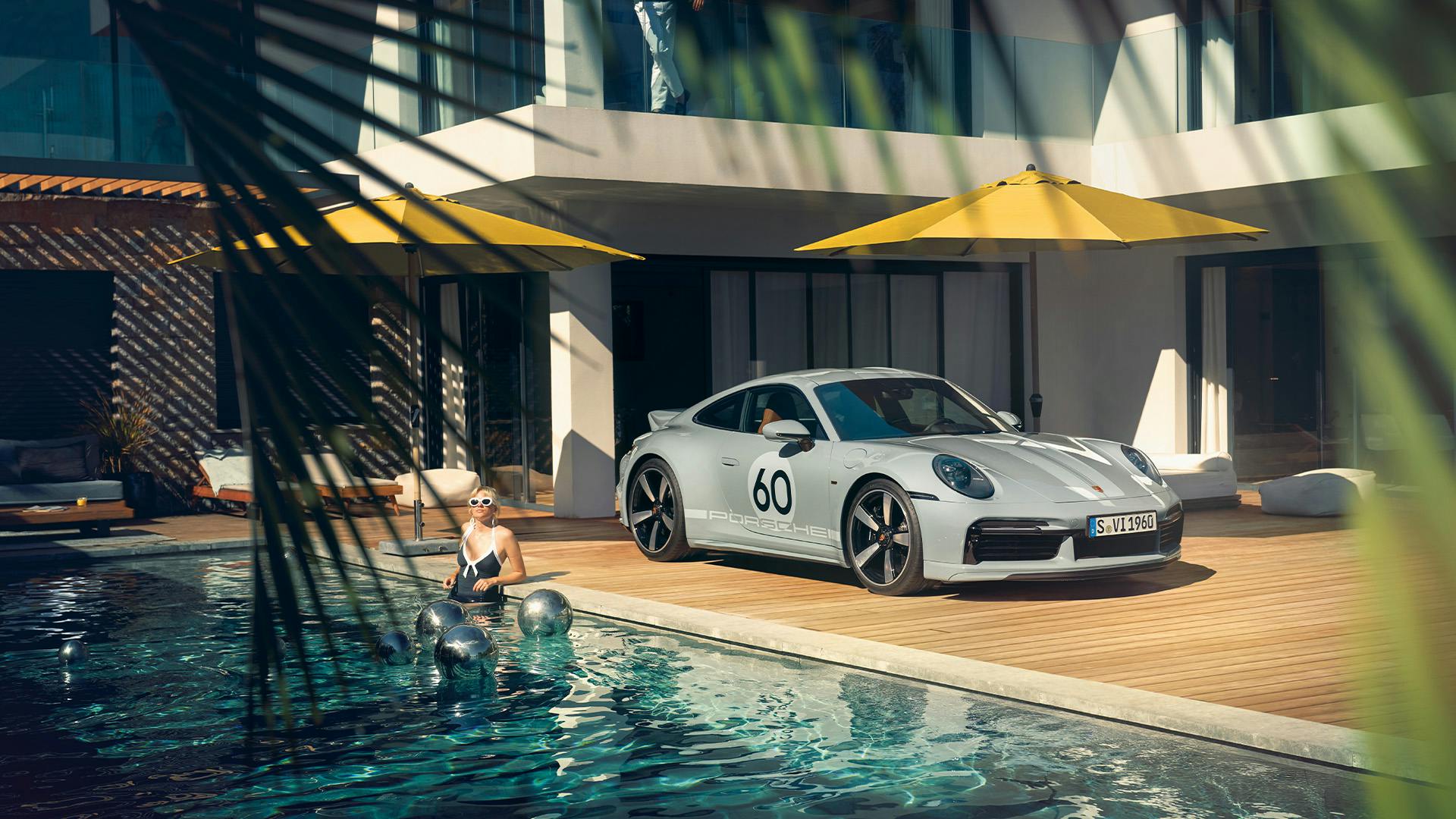 A silver Porsche on a wooden terrace in front of a modern hotel, a woman standing in the sunlight in a pool.