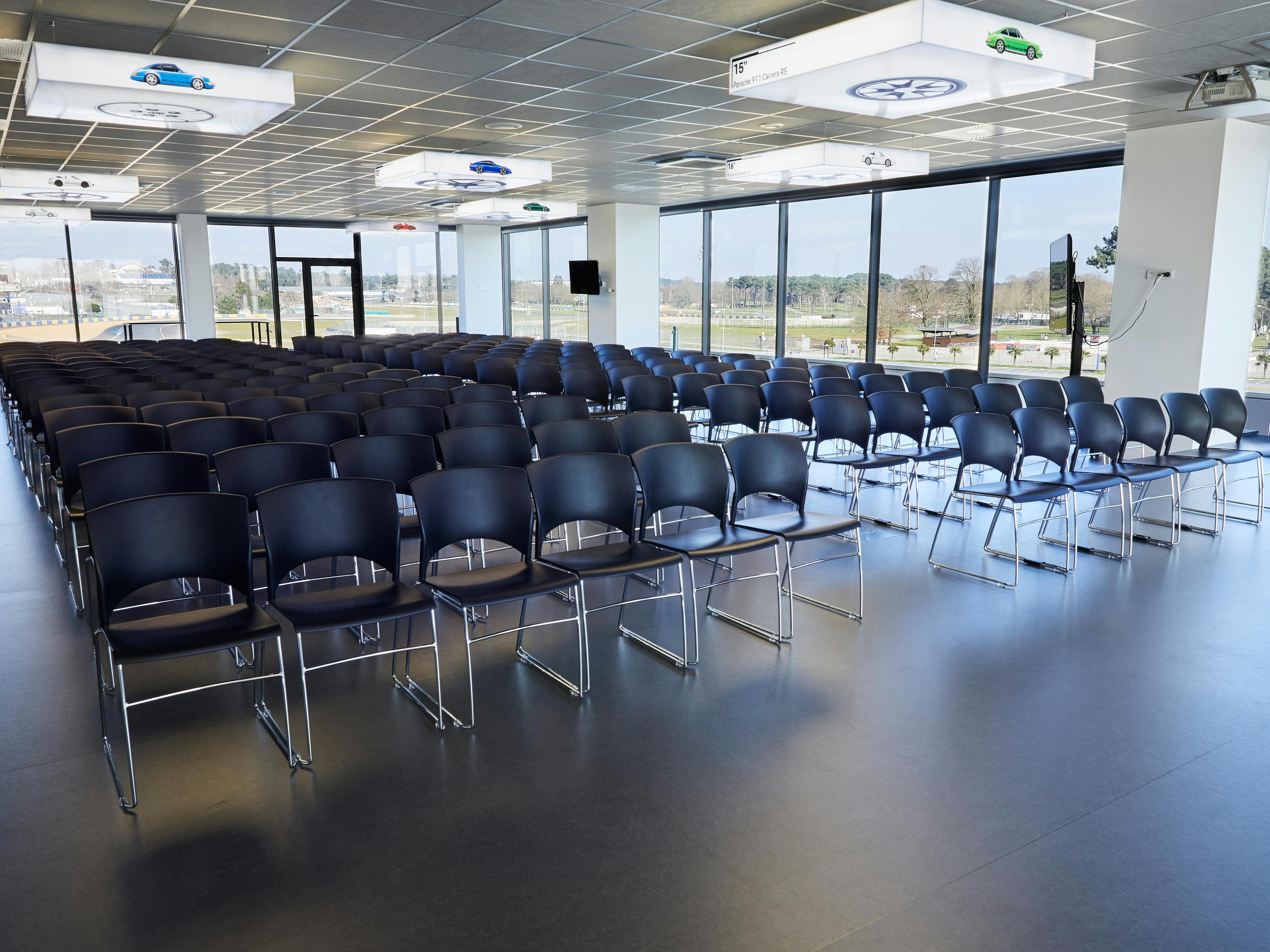 Salle 917 du Porsche Experience Centre en mode plénière, 200 chaises vides et au fond de la salle, vue sur le circuit Bugatti.