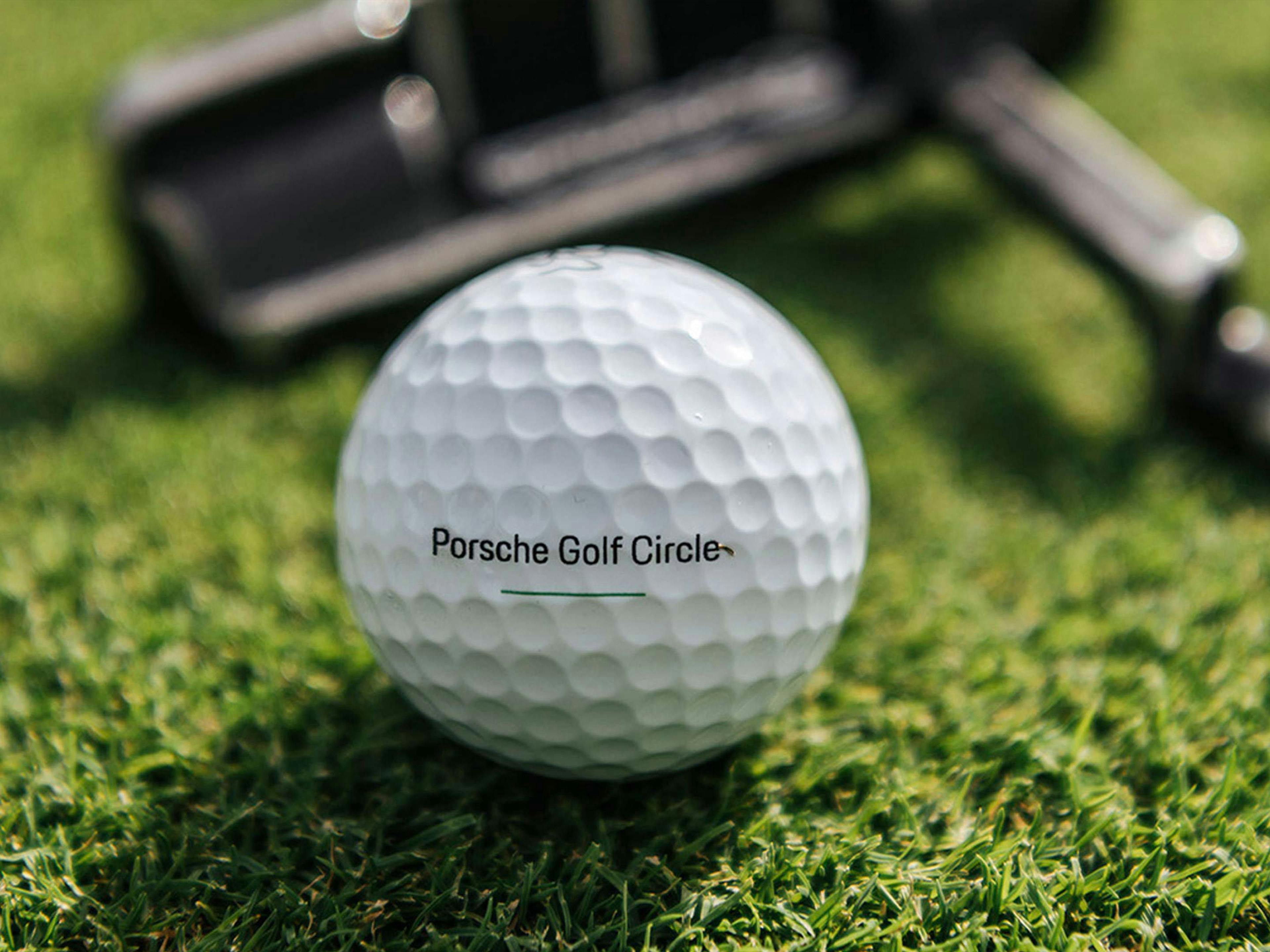 Eine Golfball mir der Aufschrift Porsche Golf Circle auf einem grünen Rasen und einem Golfschläger im Hintergrund