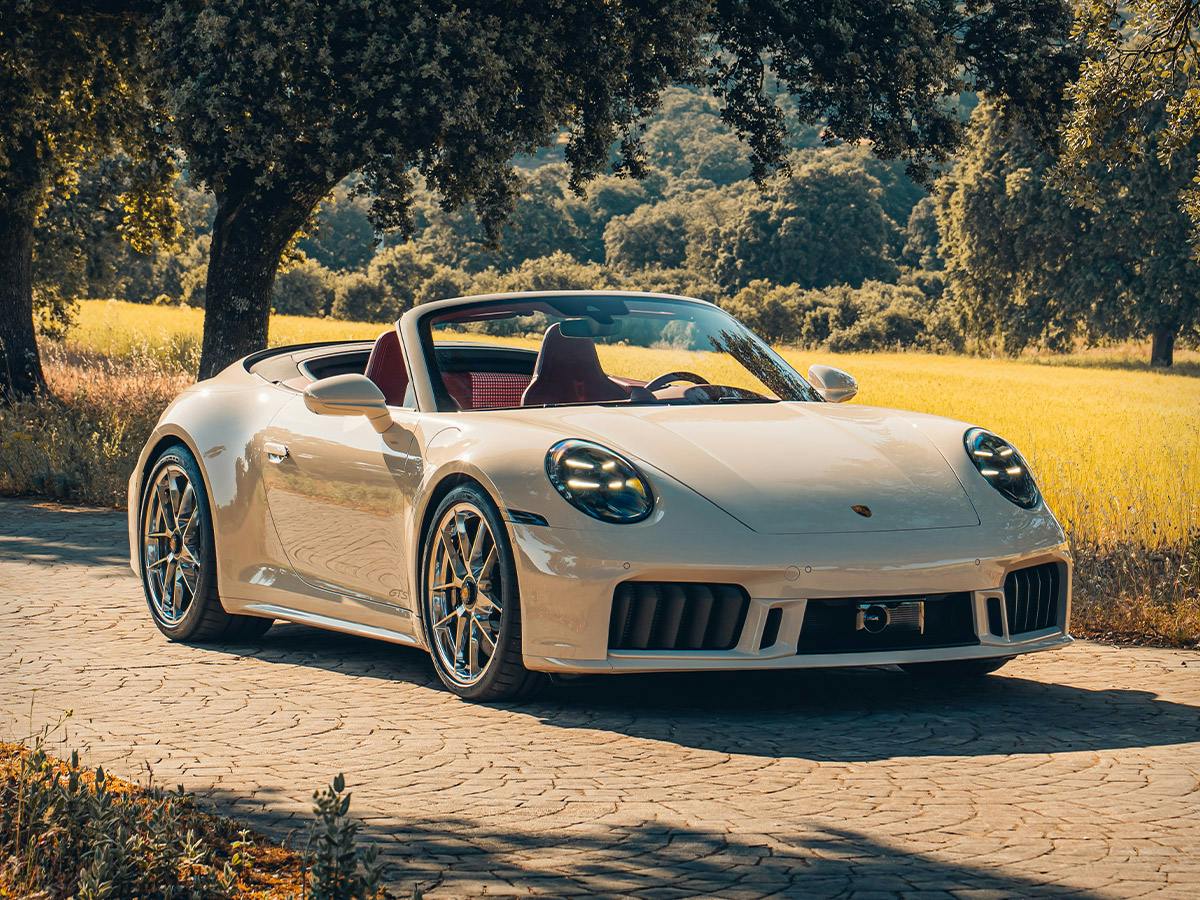 Front three‑quarter view of the 911 Carrera GTS Cabriolet in Paint to Sample Cremewhite with Neodyme accents driving on a tree‑lined country road.