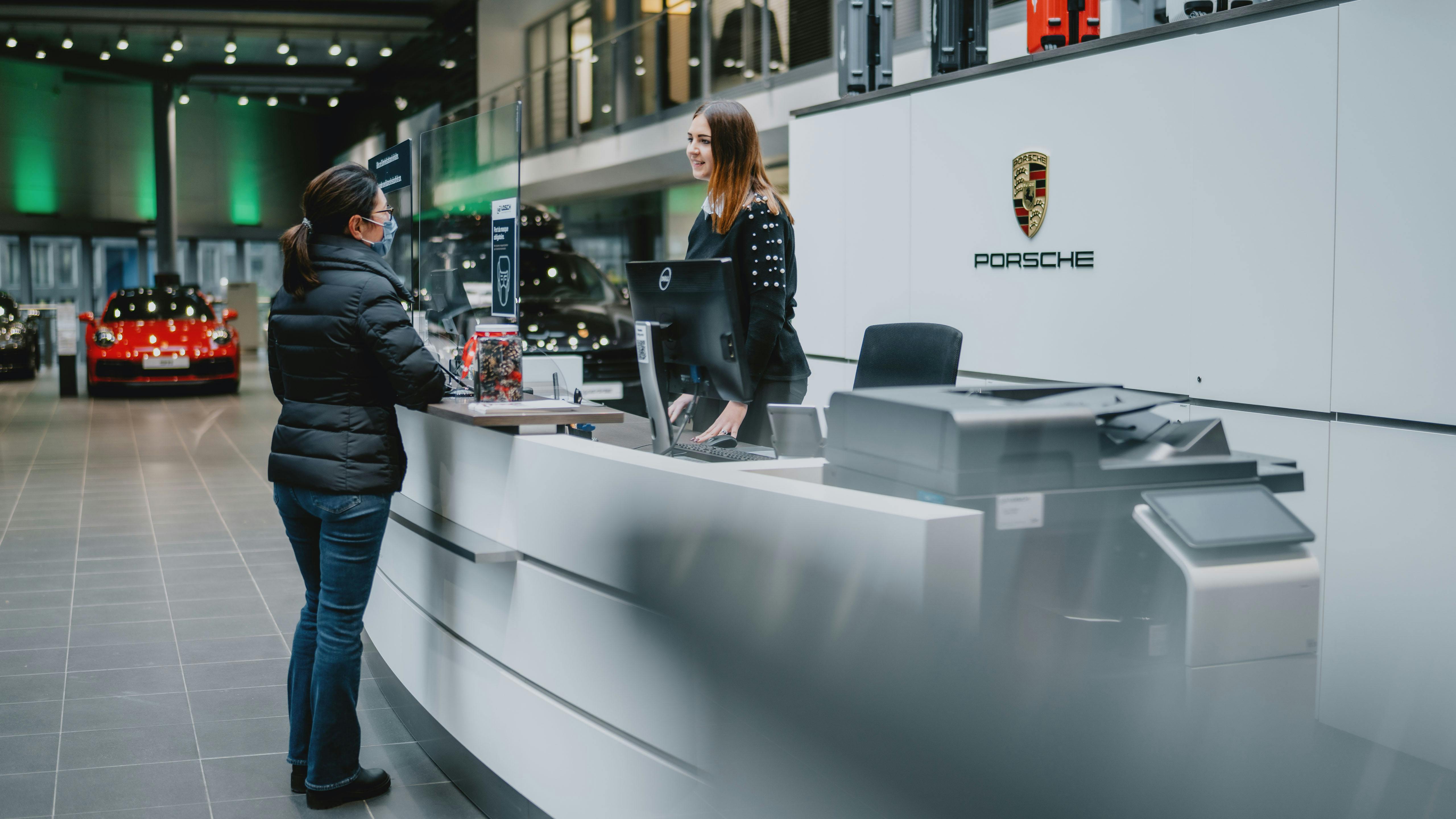 Vue latérale de la longue réception blanche au Porsche Zenter Lëtzebuerg, avec une employée de Porsche derrière le comptoir de réception en dialogue avec une cliente devant le comptoir.
