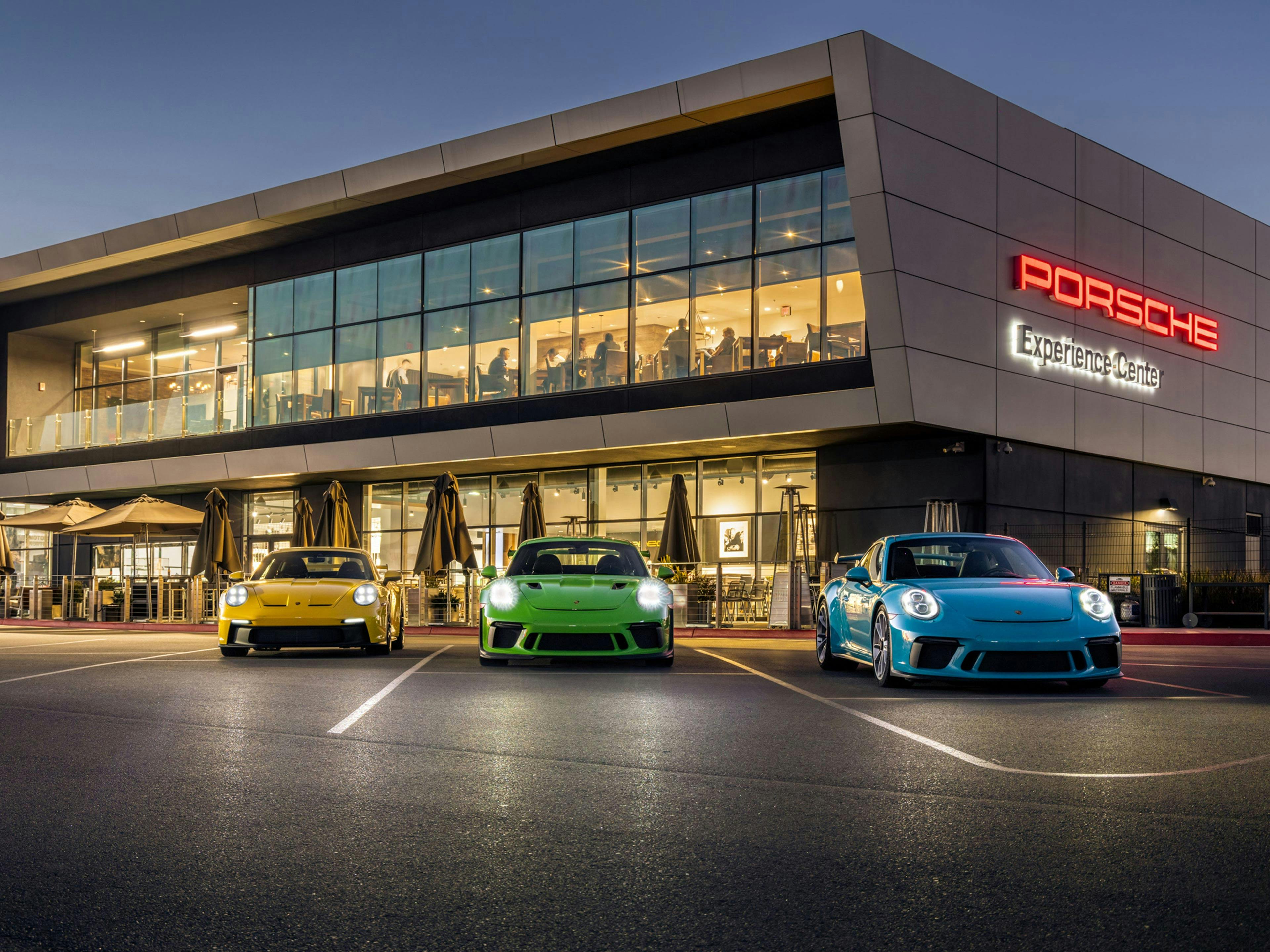 Delante del Porsche Experience Center Los Ángeles están aparcados en fila tres vehículos Porsche con el atardecer de fondo.