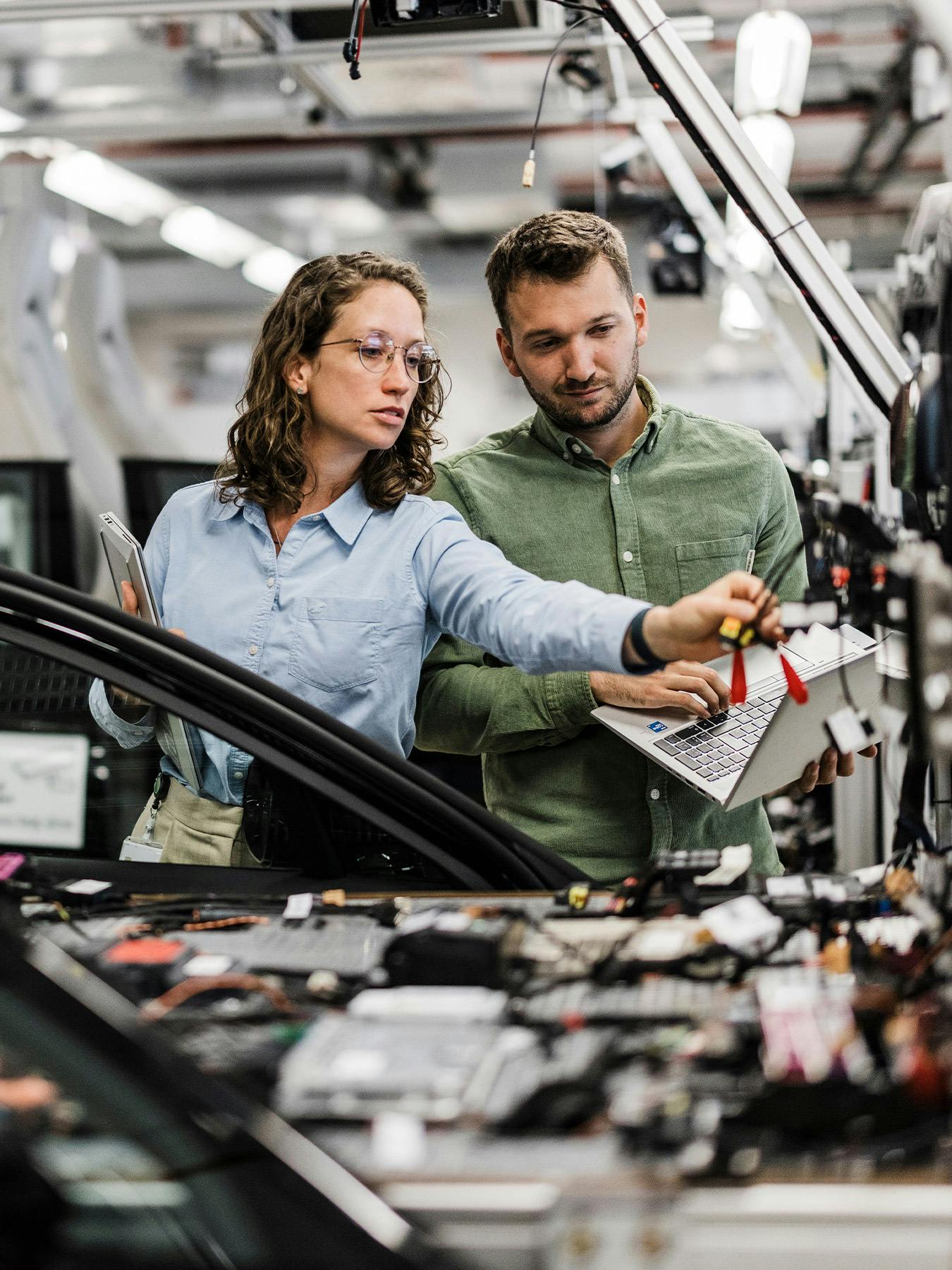 Zwei Personen arbeiten gemeinsam an einem Fahrzeug in einer modernen Produktionshalle, stehen neben einem geöffneten Auto mit sichtbaren Kabeln und Bauteilen und nutzen Werkzeuge sowie Unterlagen.