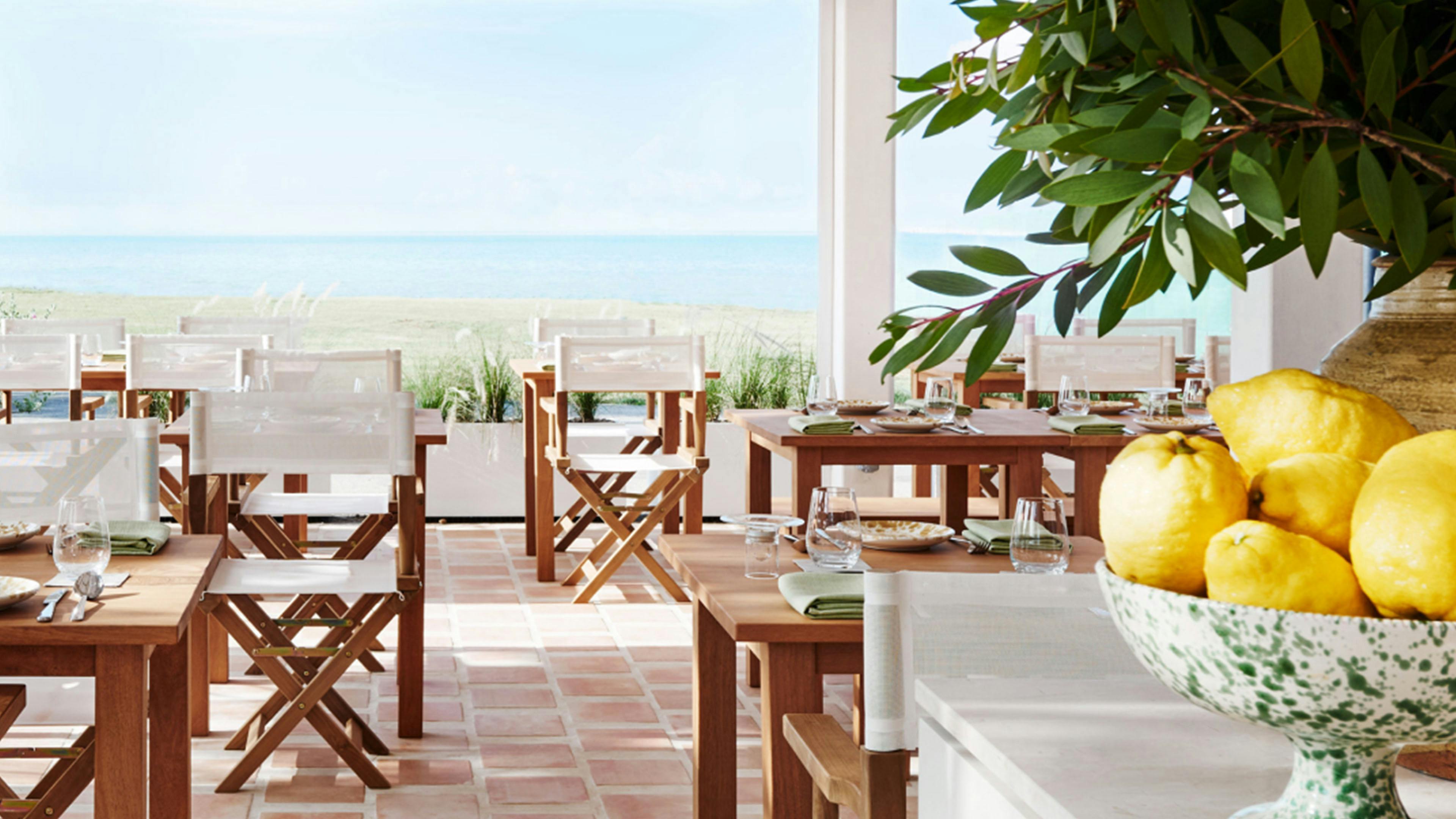 Open-air dining area with wooden tables, white chairs, terracotta floor at Fico East Coast overlooking the ocean.