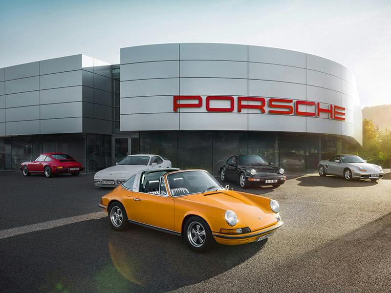 Verschiedene Porsche Modelle (vom 356 bis zum Carrera GT) auf dem Parkplatz eines Classic Partners mit gelbem 911er Carrera Cabriolet Classic im Vordergrund.