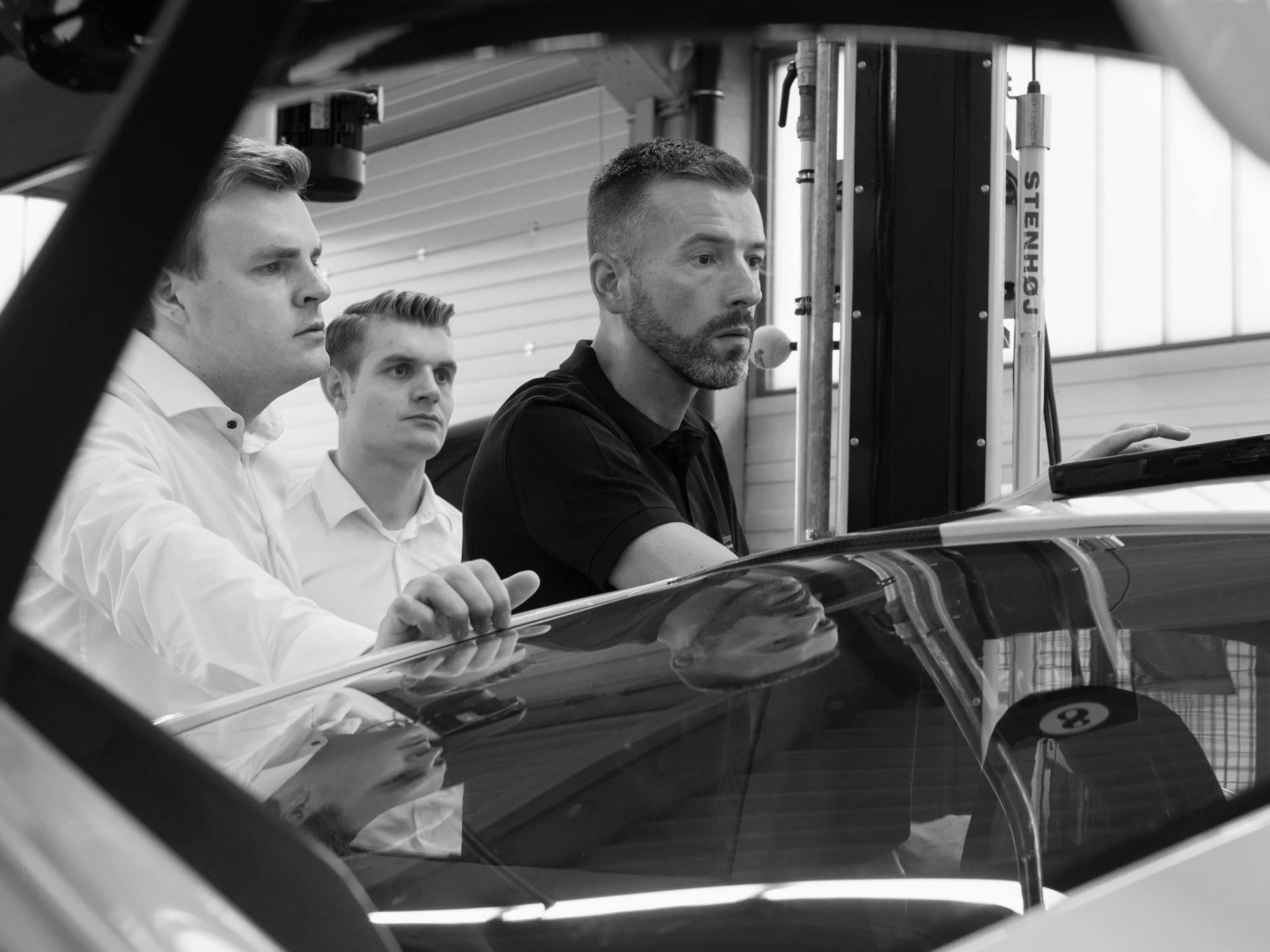 Black-and-white photo: three Porsche employees in a workshop look critically at a display.