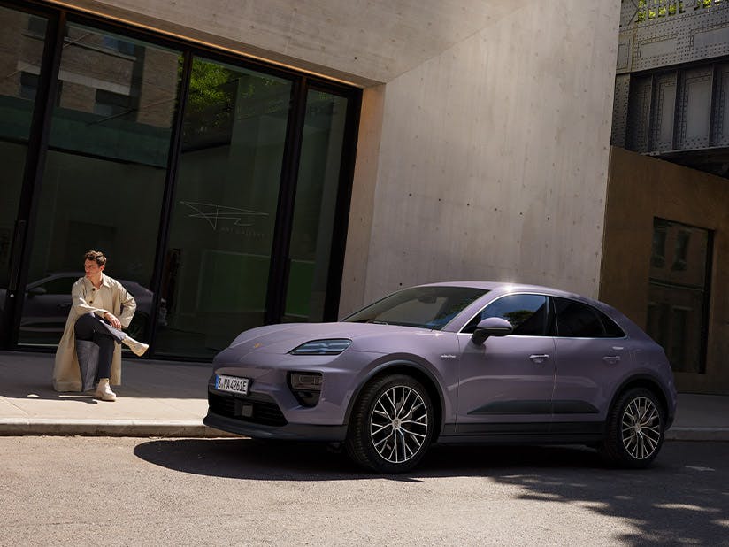Porsche Macan steht vor einem modernen Betongebäude. Eine Person sitzt links neben dem Fahrzeug auf einer niedrigen Stufe am Gebäude.