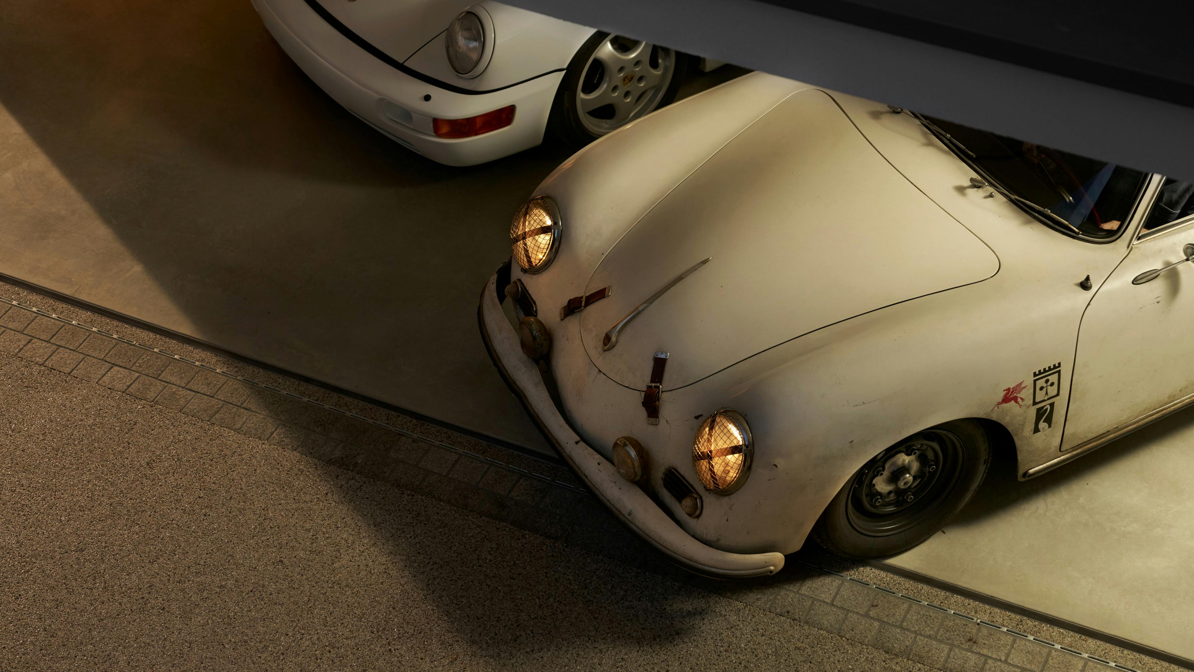 A white classic Porsche car standing in a garage with its headlights on.
