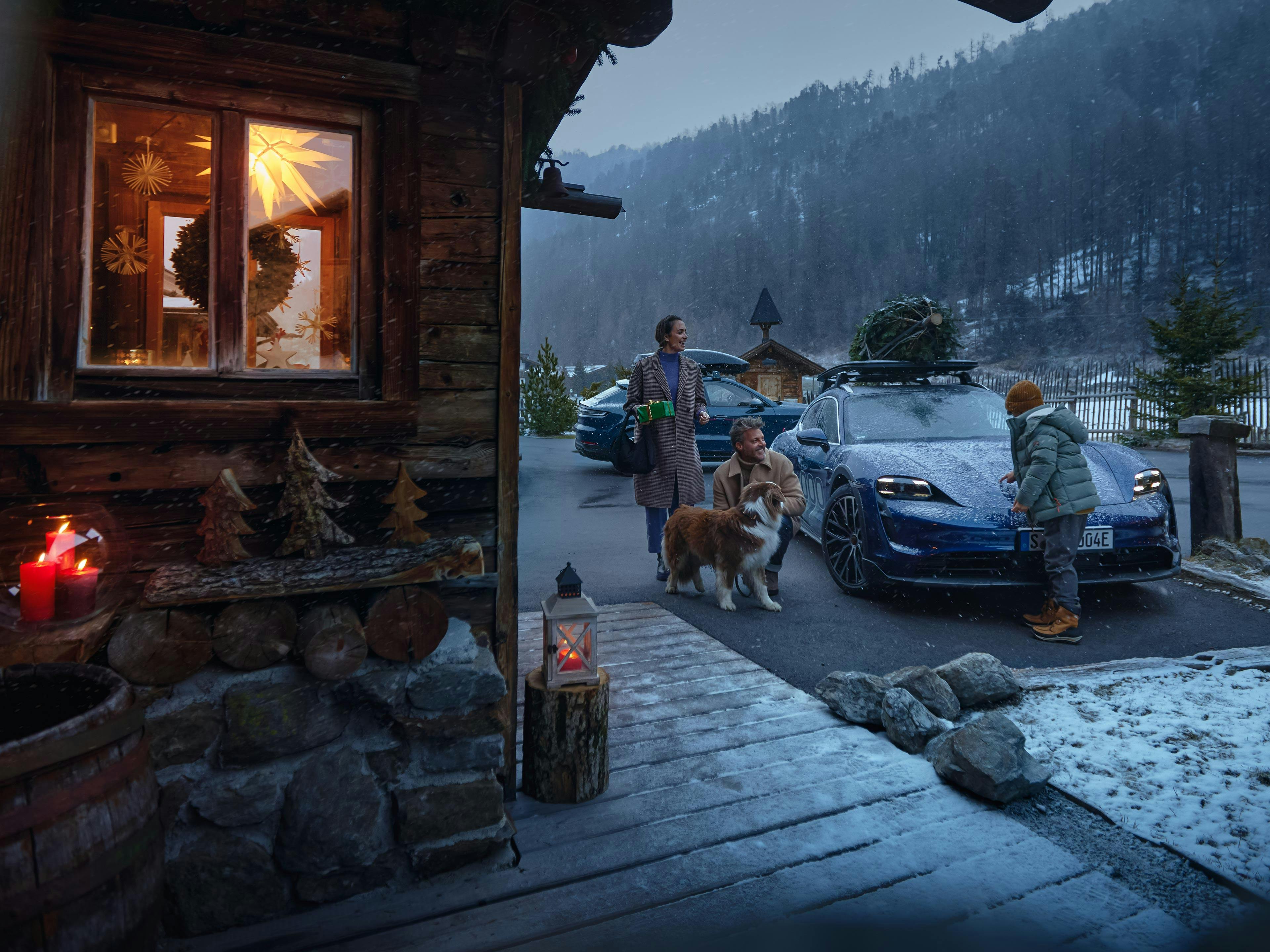 Eine Familie mit Hund bringt einen Tannenbaum mit einem Porsche Taycan in das winterliche Ferienhaus in den Bergen.