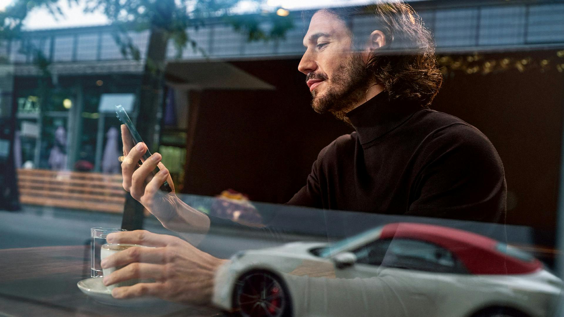 Un hombre se sienta en la ventana y mira su teléfono móvil. Un coche Porsche se refleja en la ventana.