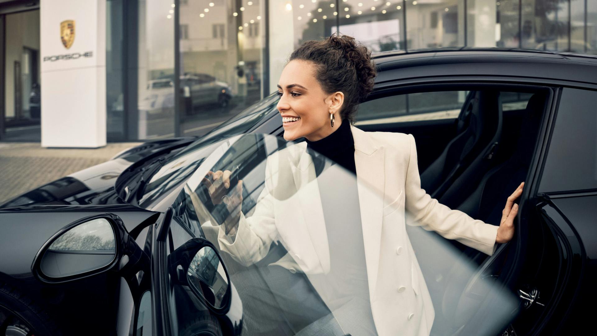 A smiling woman in a white blazer opening the door of a black Porsche, with a Porsche dealership visible in the background.