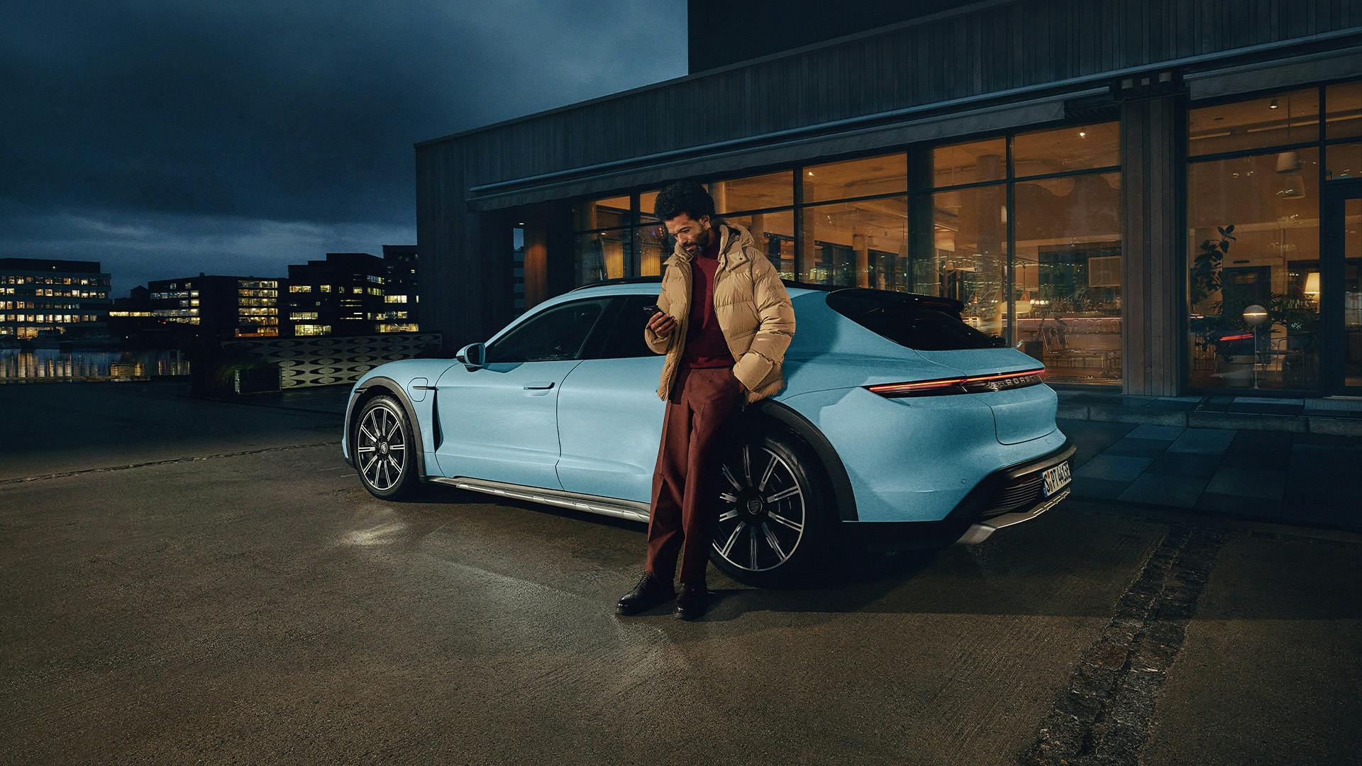 Ein Mann lehnt in der Nacht an einem blauen Porsche und schaut auf sein Handy.