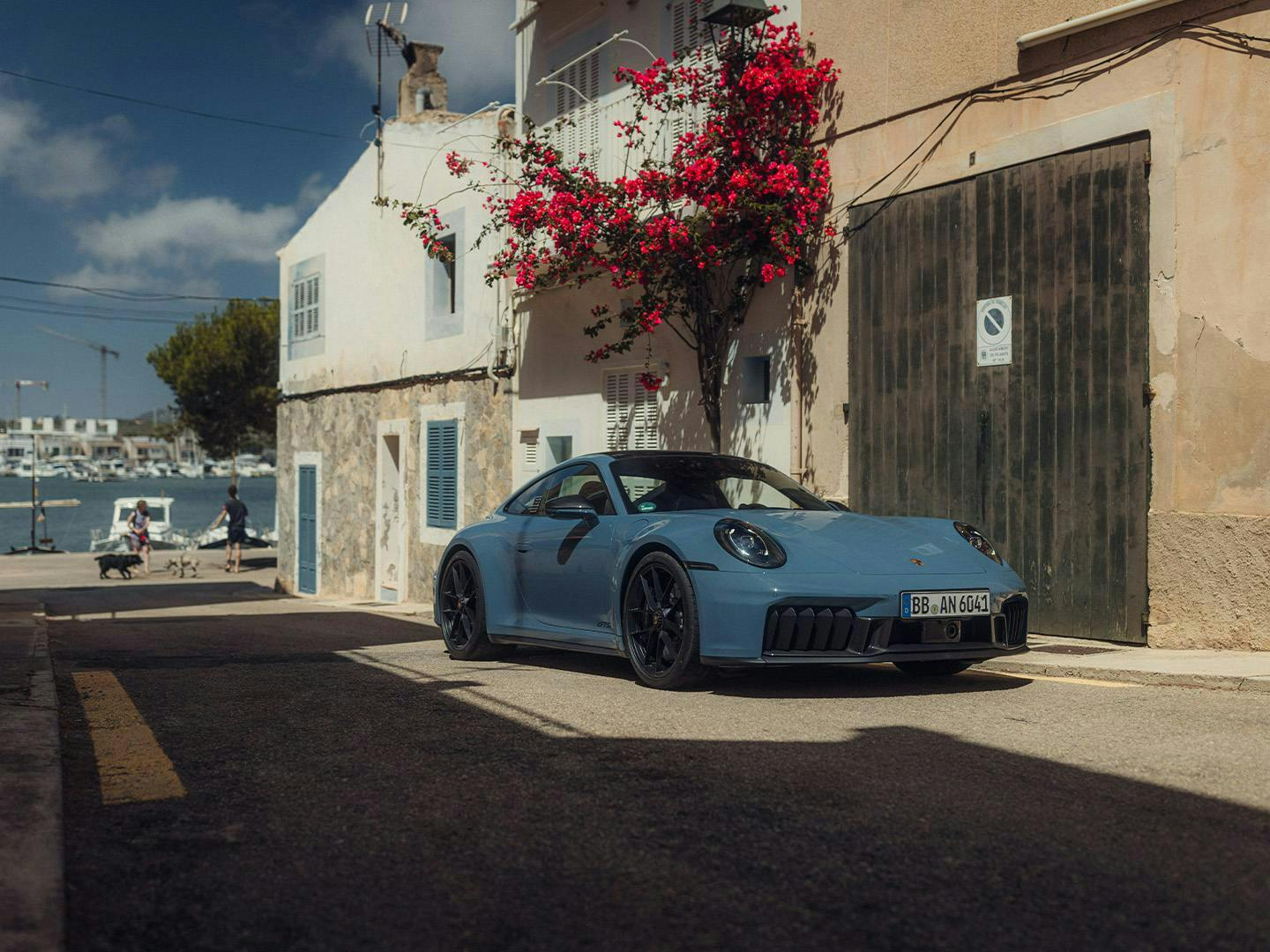 Light blue Porsche 911 in front of a house facade with pink flowers; the sea is visible in the background of the alley.