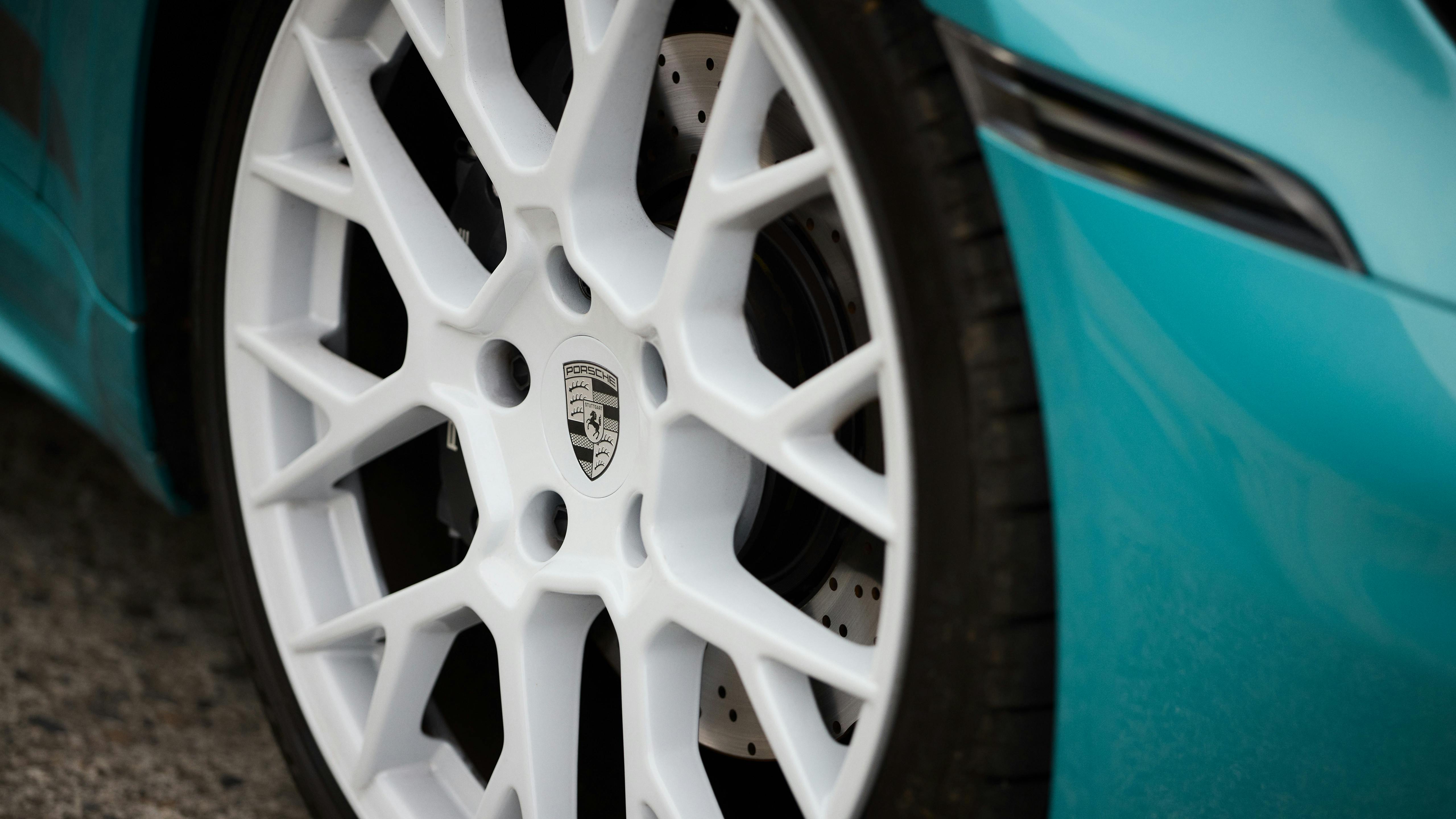 Close-up of a white multi-spoke alloy wheel on a turquoise Porsche, showing brake disc and tire edge.
