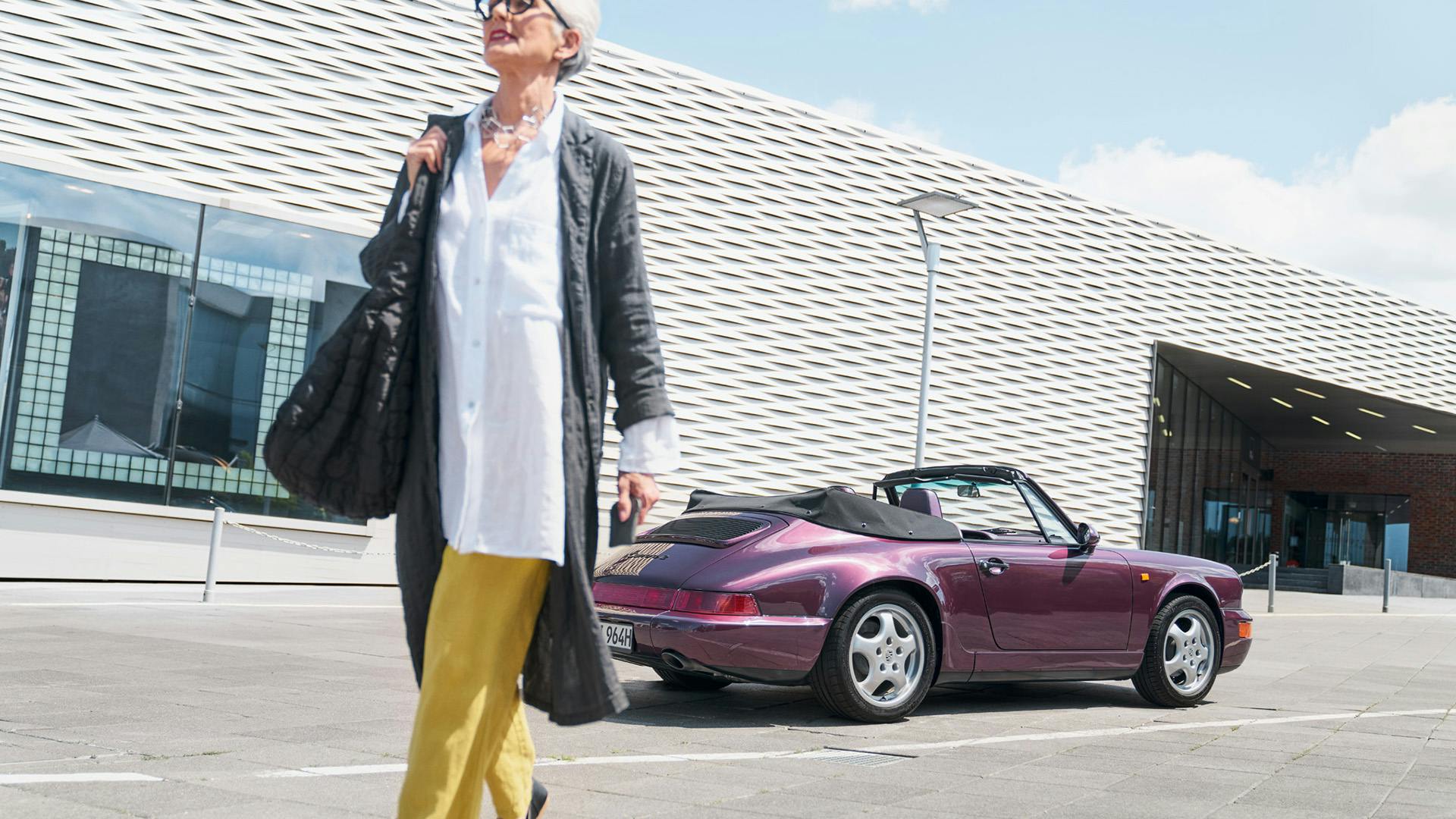 Street scene: side view of a Porsche 911 (Type 964) in aubergine color, parked in front of a building with a woman passing by.