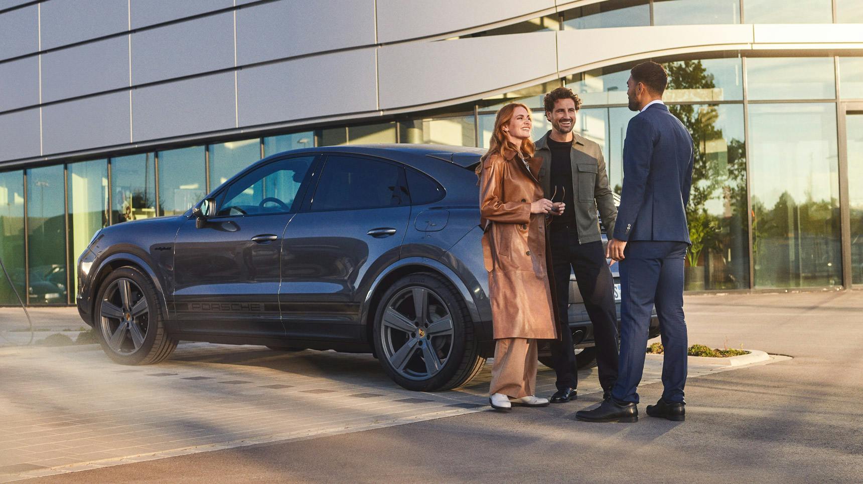 A sleek gray Porsche SUV parked outside a Porsche showroom, with three people engaged in conversation nearby.