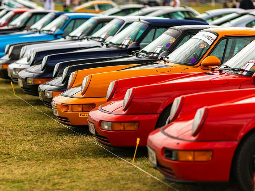 A row of classic Porsche 911s in various colours parked on grass at a Porsche car show.