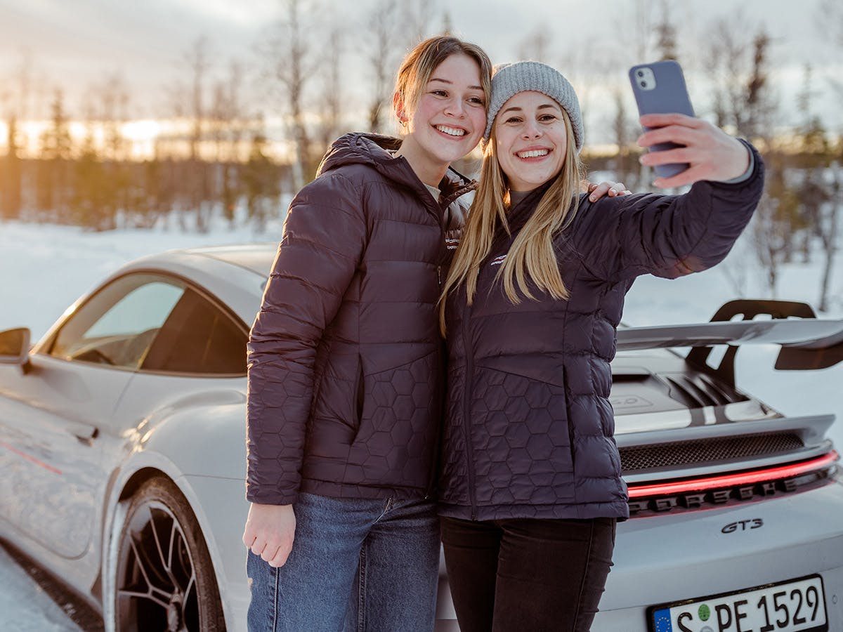 Dos participantes de Porsche Ice Experience capturan sus recuerdos con un selfie delante del vehículo y con un fondo nevado.