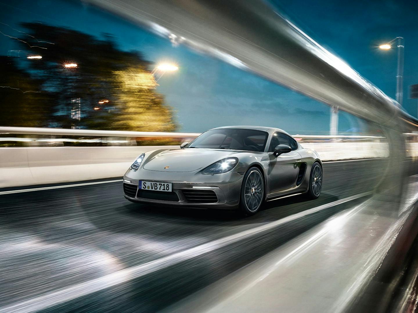 Silberner Porsche 718 Cayman fährt bei Nacht auf einer beleuchteten Straße, dynamische Bewegung mit unscharfem Hintergrund.