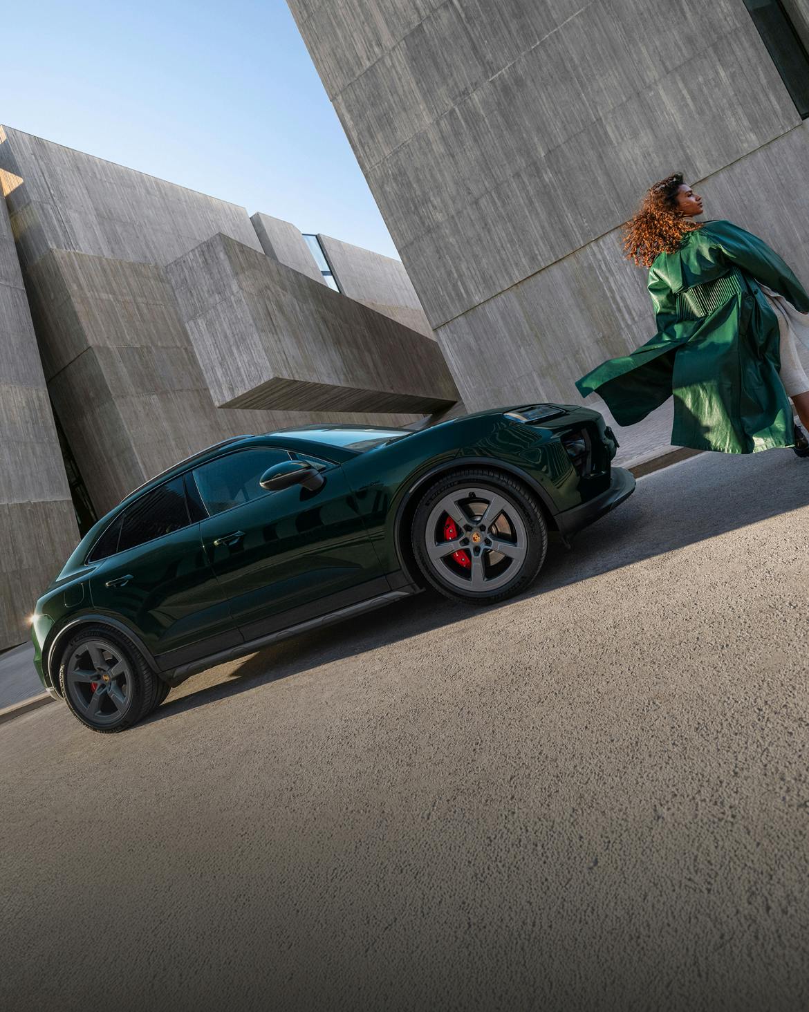A Porsche Macan 4S in Oak Green Metallic stands in front of concrete walls. A woman in a green coat walks by.