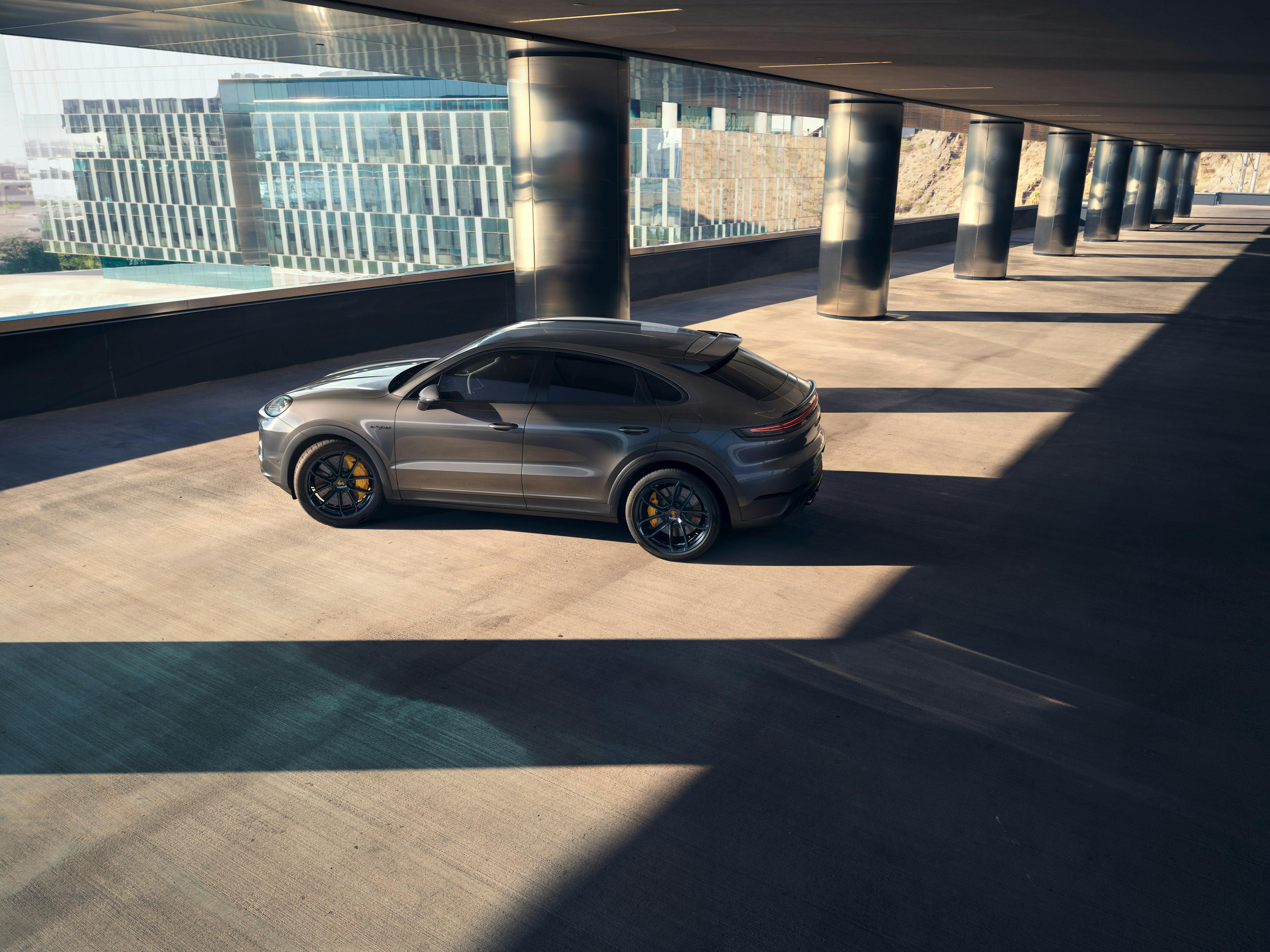 Ein dunkelgrauer SUV (Cayenne)  sportlichem Design steht in einem modernen, offenen Parkhaus. Durch die Säulen und die tiefstehende Sonne entstehen lange Schatten auf dem Betonboden. Im Hintergrund sind moderne Glasgebäude sichtbar.