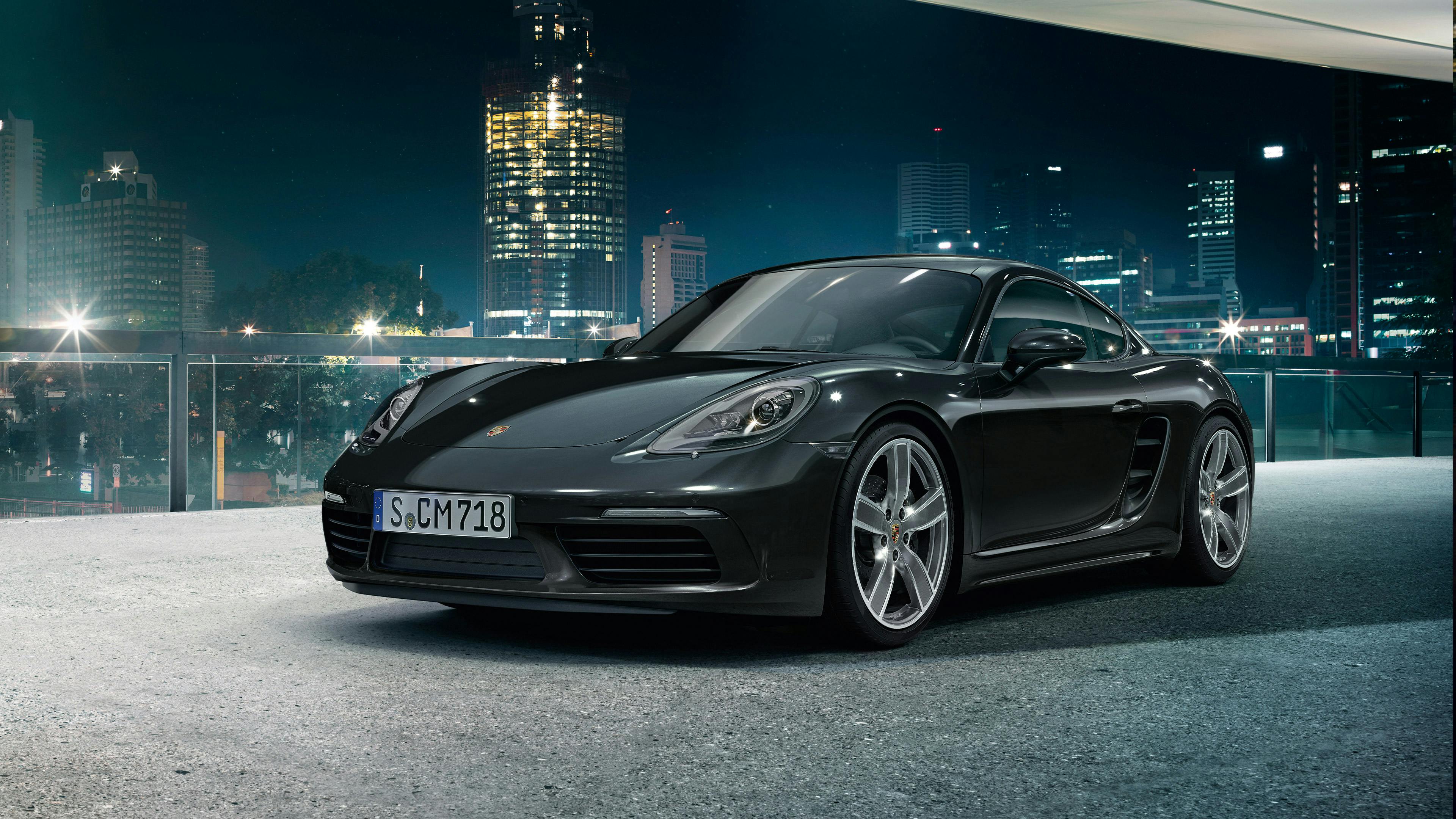 Une Porsche 718 Cayman noire est garée dans un bâtiment moderne avec une ligne d'horizon de nuit.