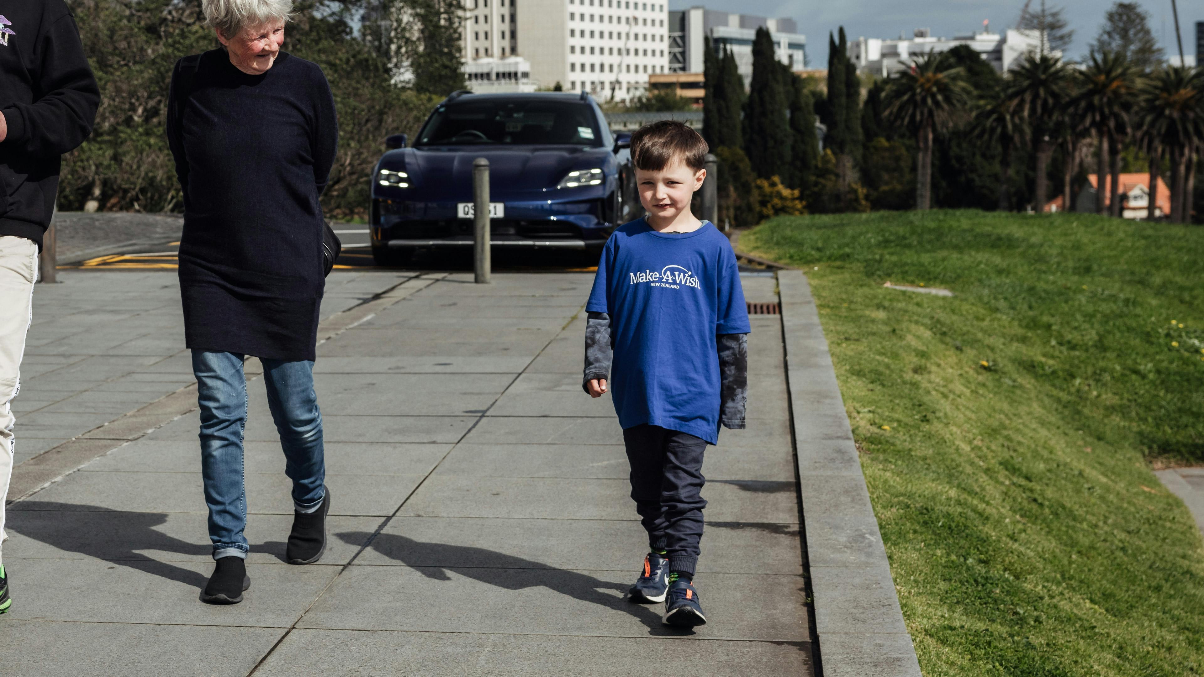 A smiling child wearing a Make-A-Wish shirt stands next to his nana in a sunny outdoor setting. The child looks excited and happy.