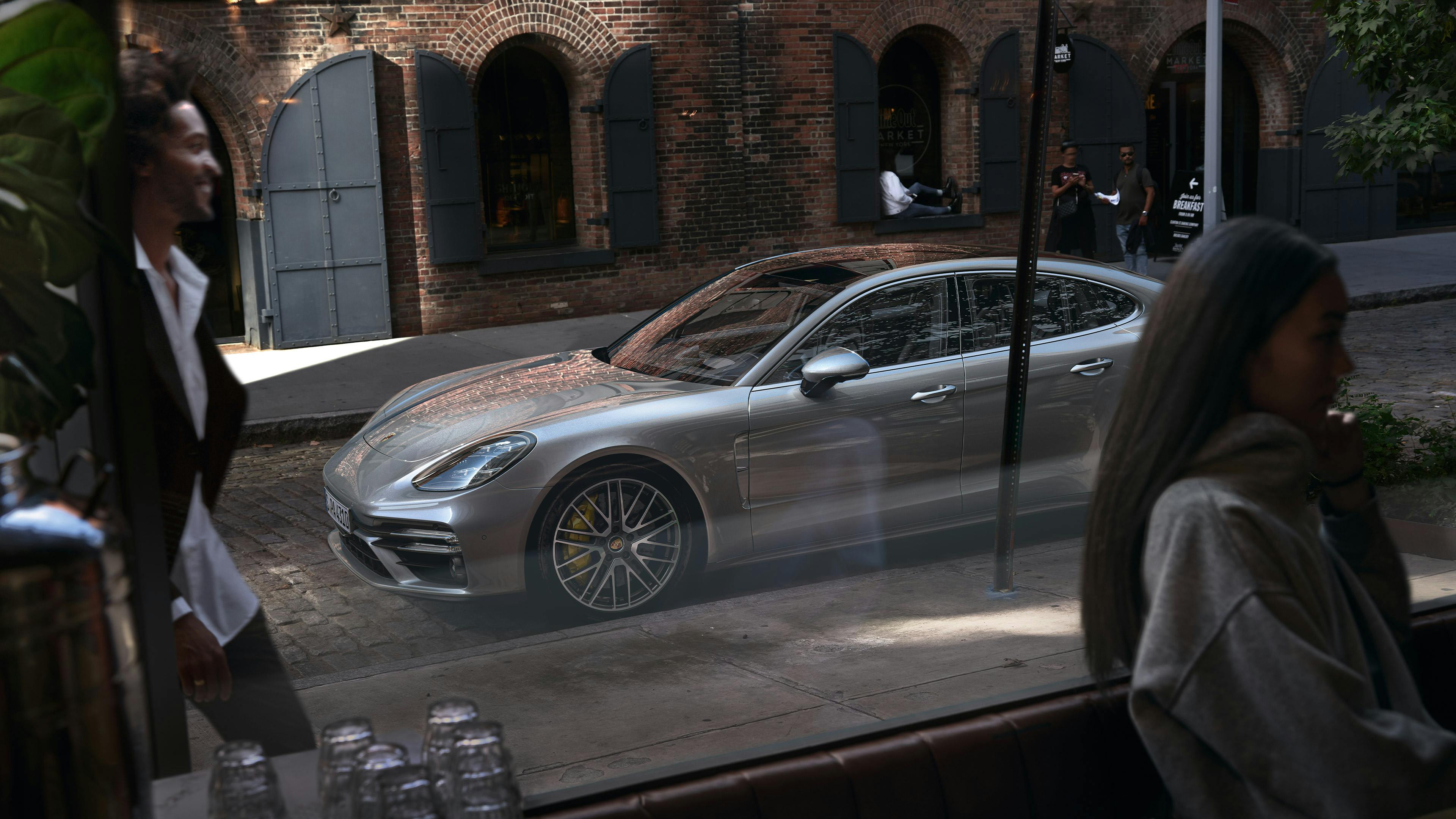 Die Kamera blickt durch die Scheibe eines Cafés. Draußen im Fokus steht ein silberner Porsche Panamera vor einem Backsteingebäude.