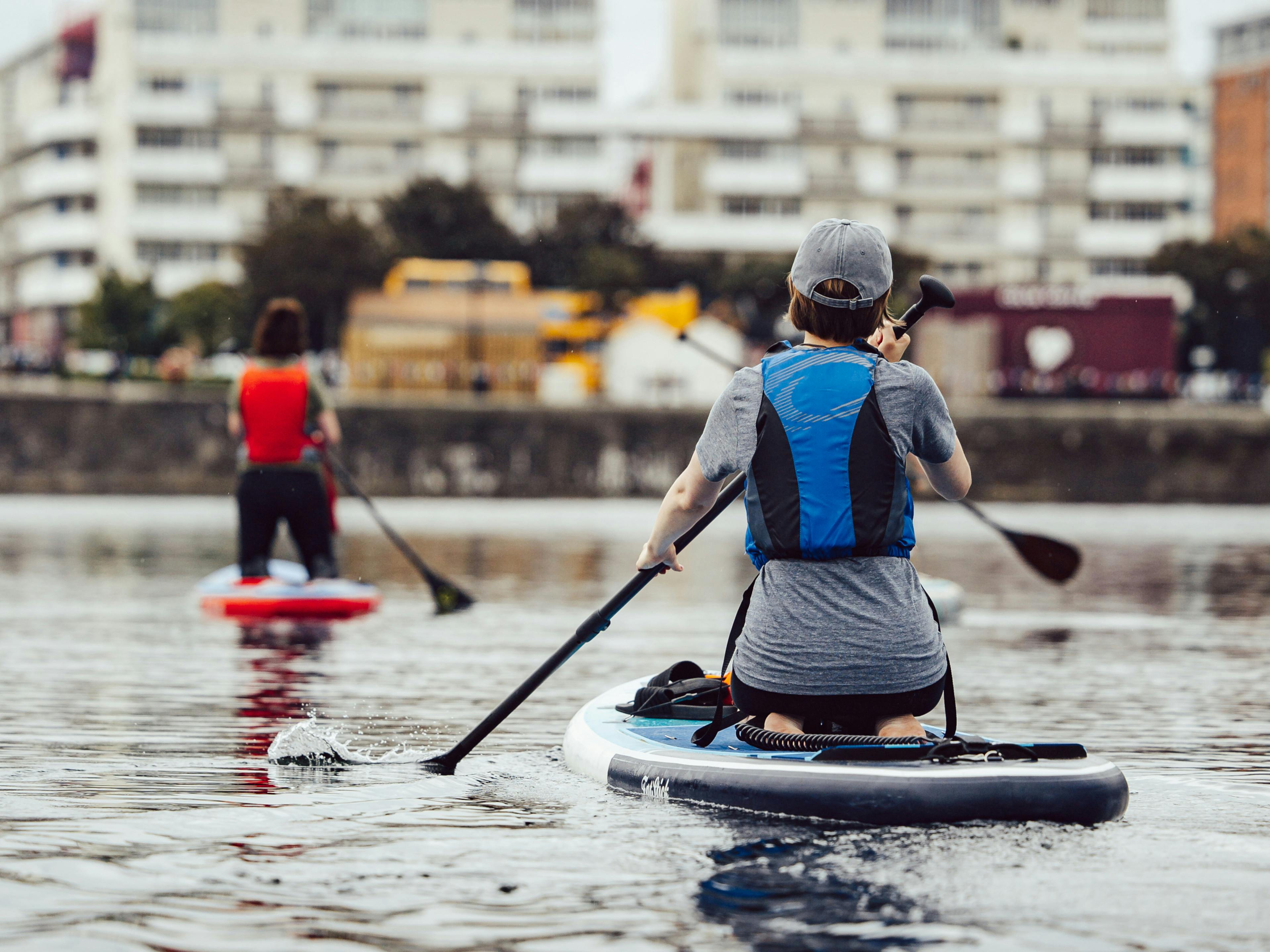 Two people are sat on separate paddleboards floating along a river, one is wearing a blue lifejacket, the other is wearing a red lifejacket.​