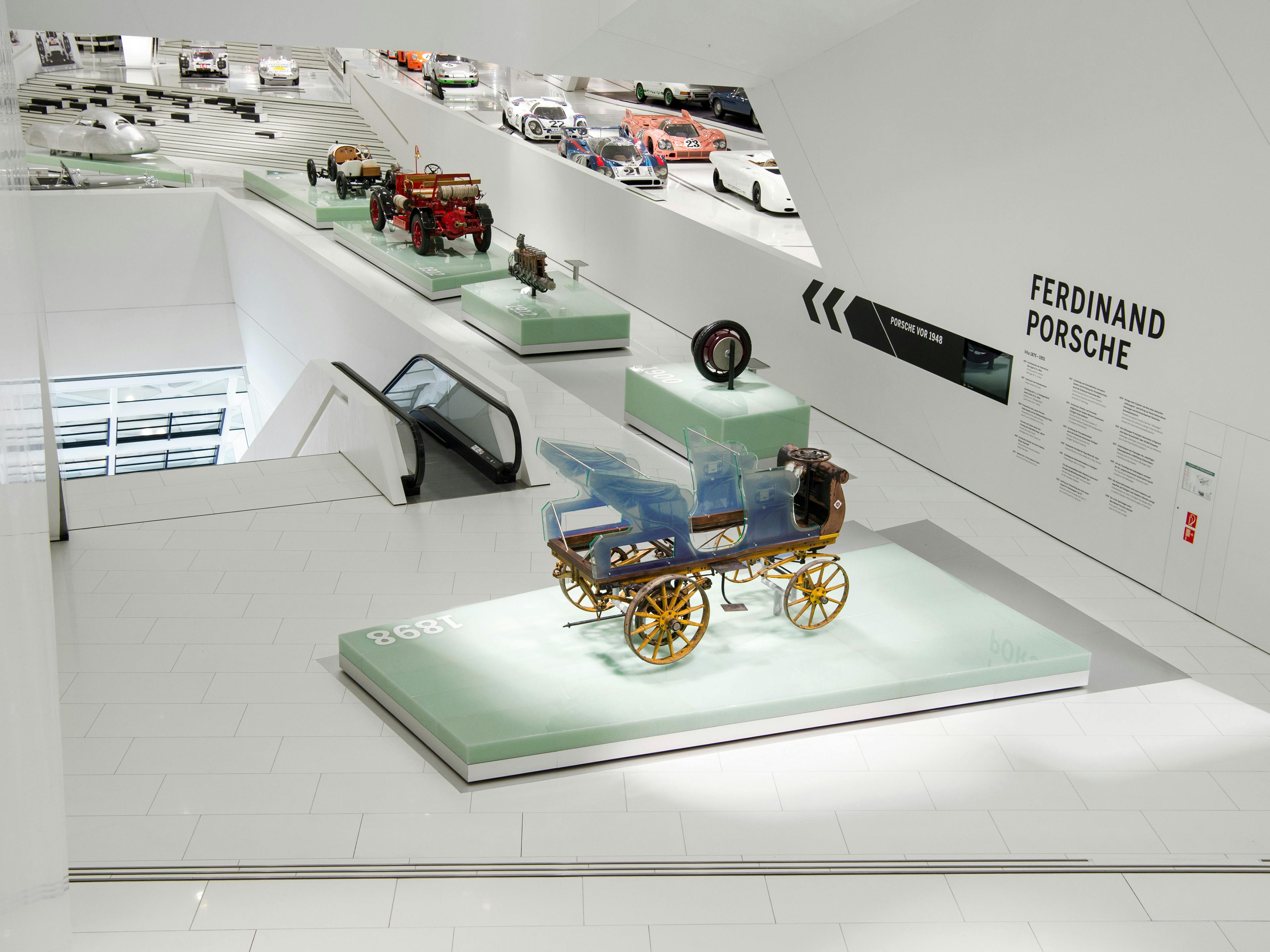 Historic Porsche vehicles and early automotive artifacts displayed on illuminated platforms in the Porsche Museum, with "Ferdinand Porsche" exhibition signage visible.