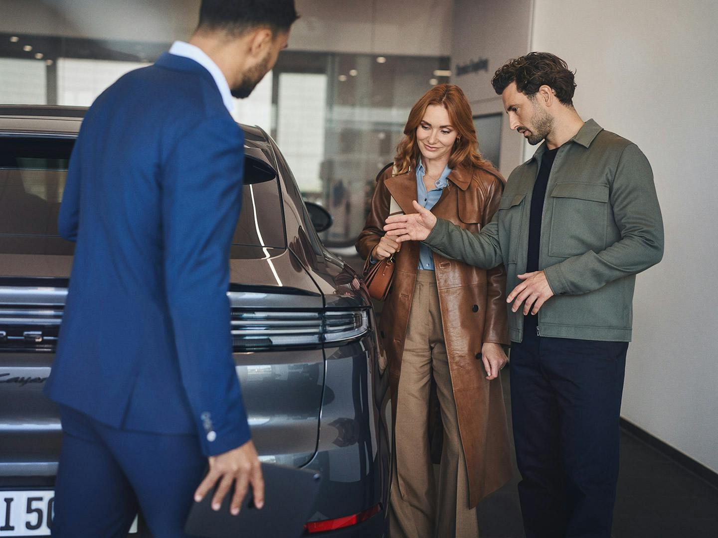 Porsche employee in a blue suit presents a Porsche car to a couple in the showroom.